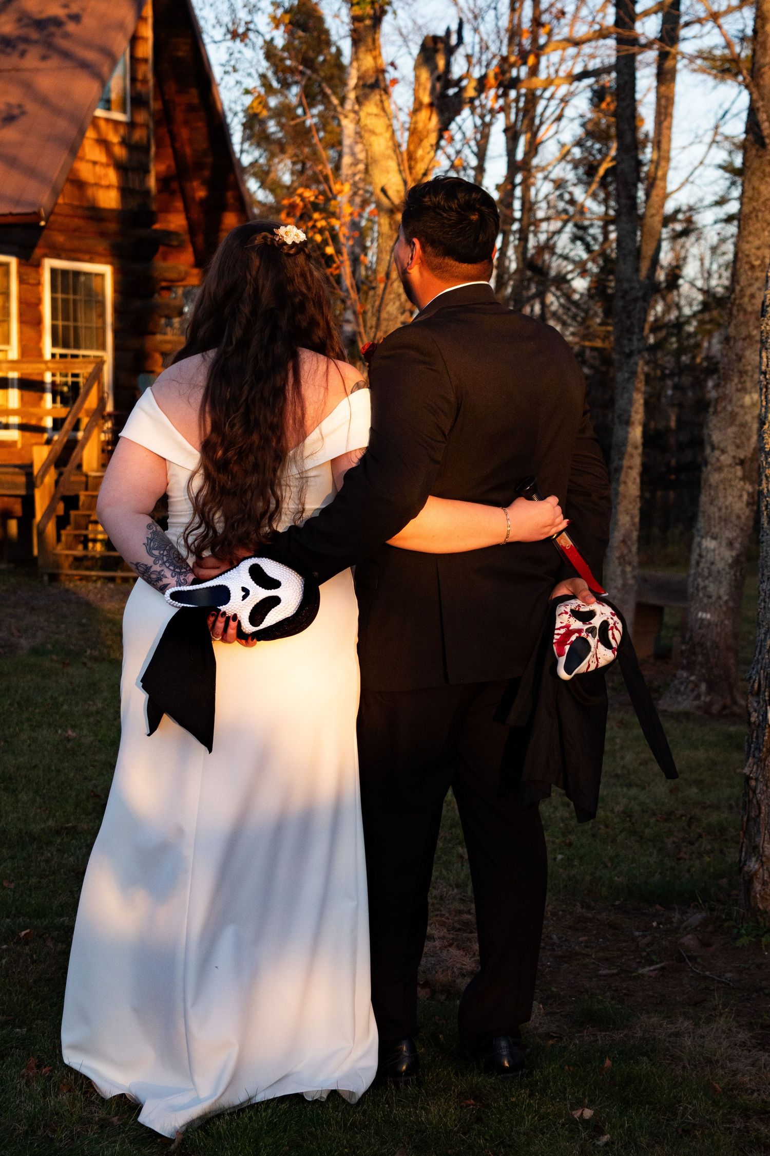 Bride and groom holding Scream Halloween masks walking away from rustic cabin venue.