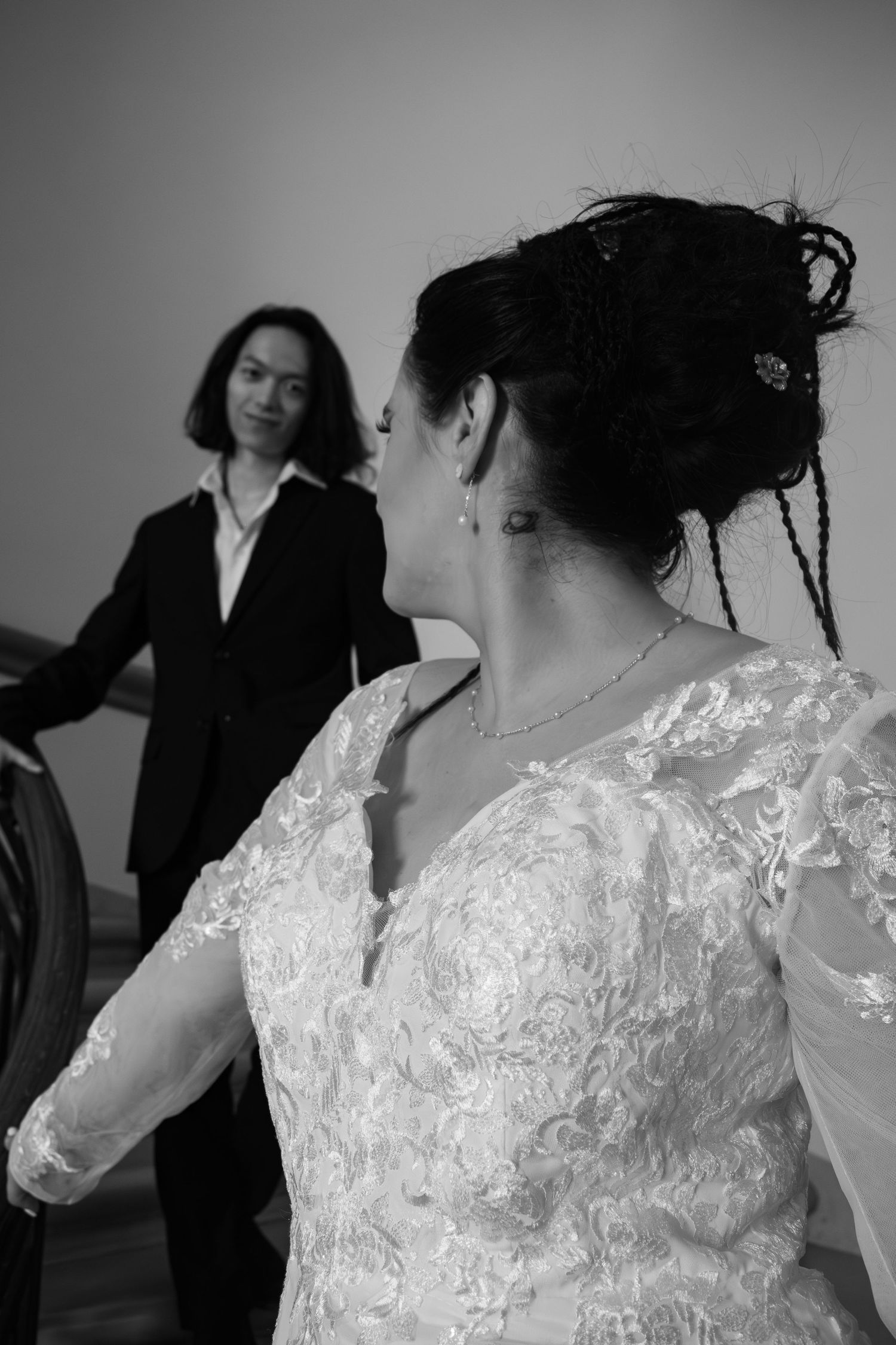 Black and white photo of bride in lace wedding gown turning toward smiling guest on staircase.