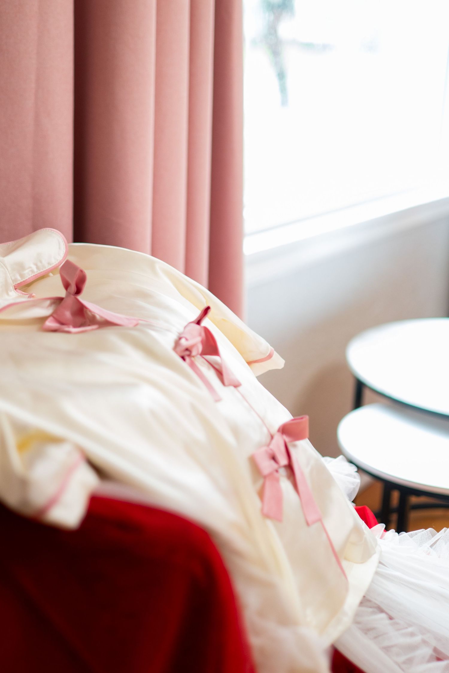 Elegant cream dress with pink satin bows displayed on a red chair near pink curtains and a window.
