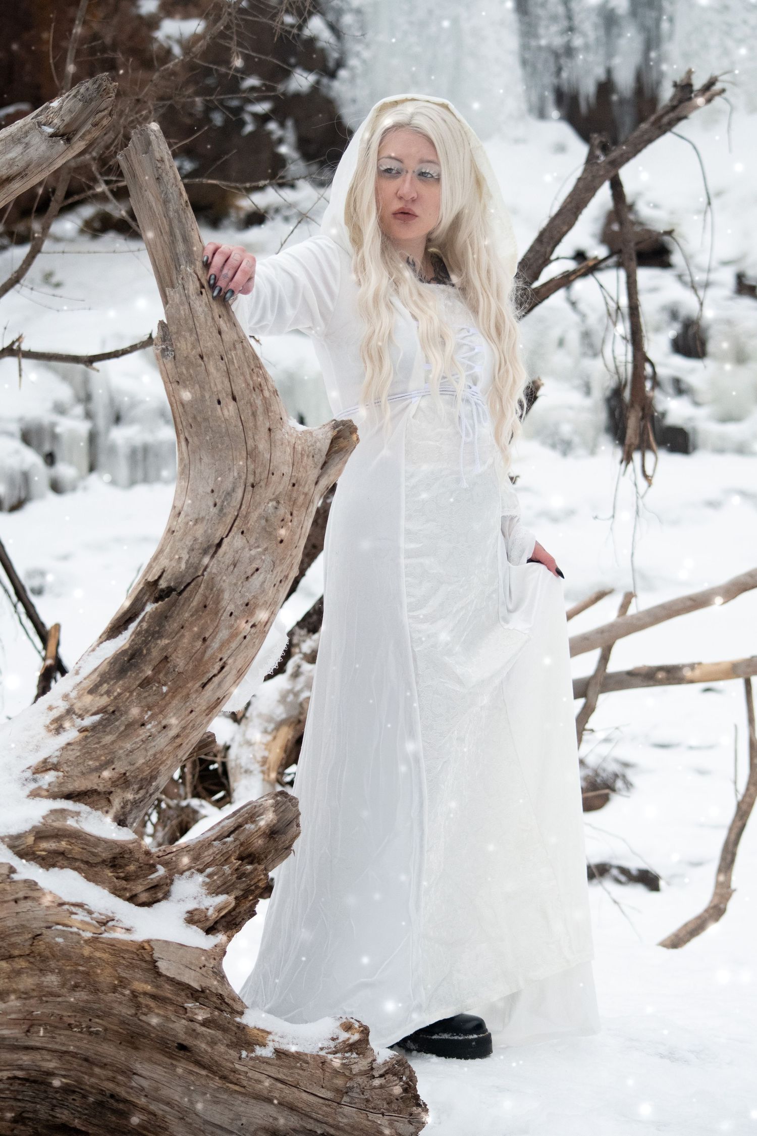 Blonde woman in white hooded medieval gown standing by driftwood in snowy landscape.