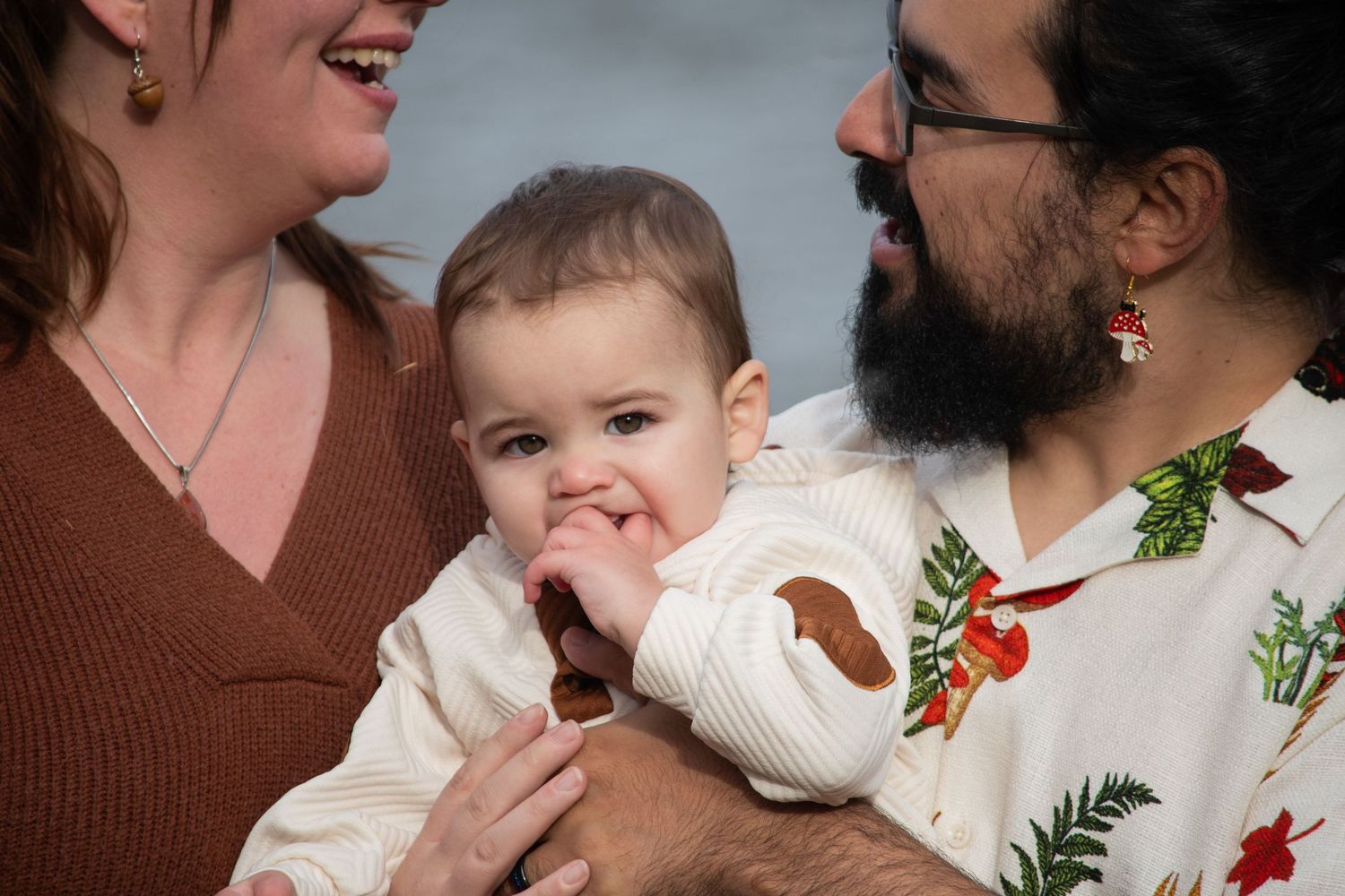 Parents smiling at baby chewing fingers, father in mushroom print shirt outdoors.