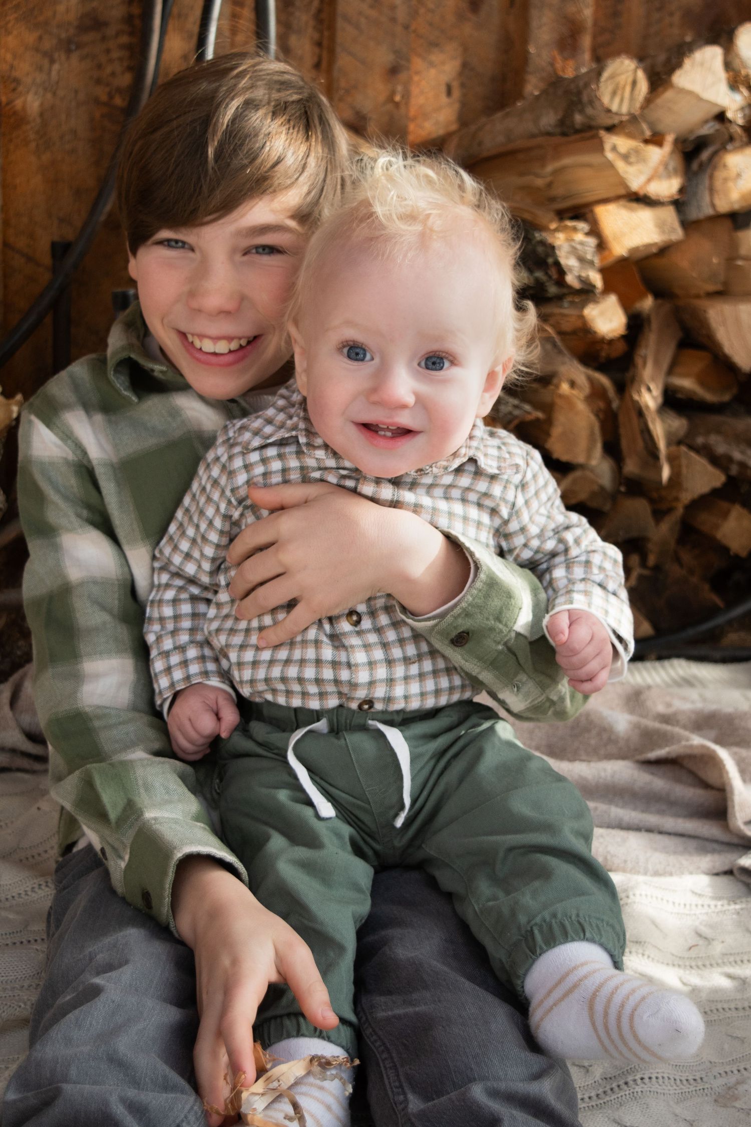 Older brother hugging smiling baby sibling in front of rustic stacked firewood background.