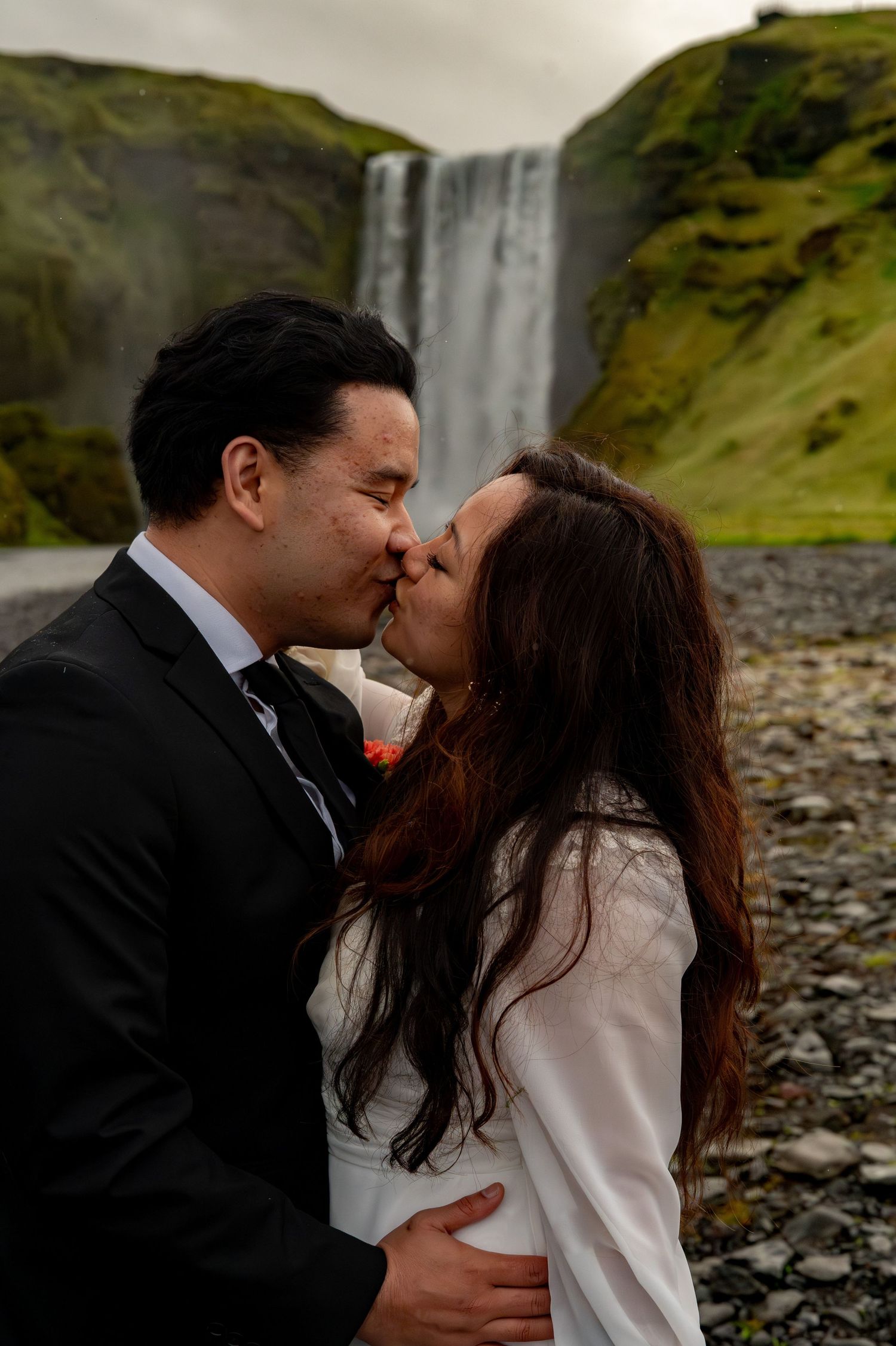 Couple sharing a romantic kiss in wedding attire with Skogafoss waterfall in Iceland as a stunning backdrop.