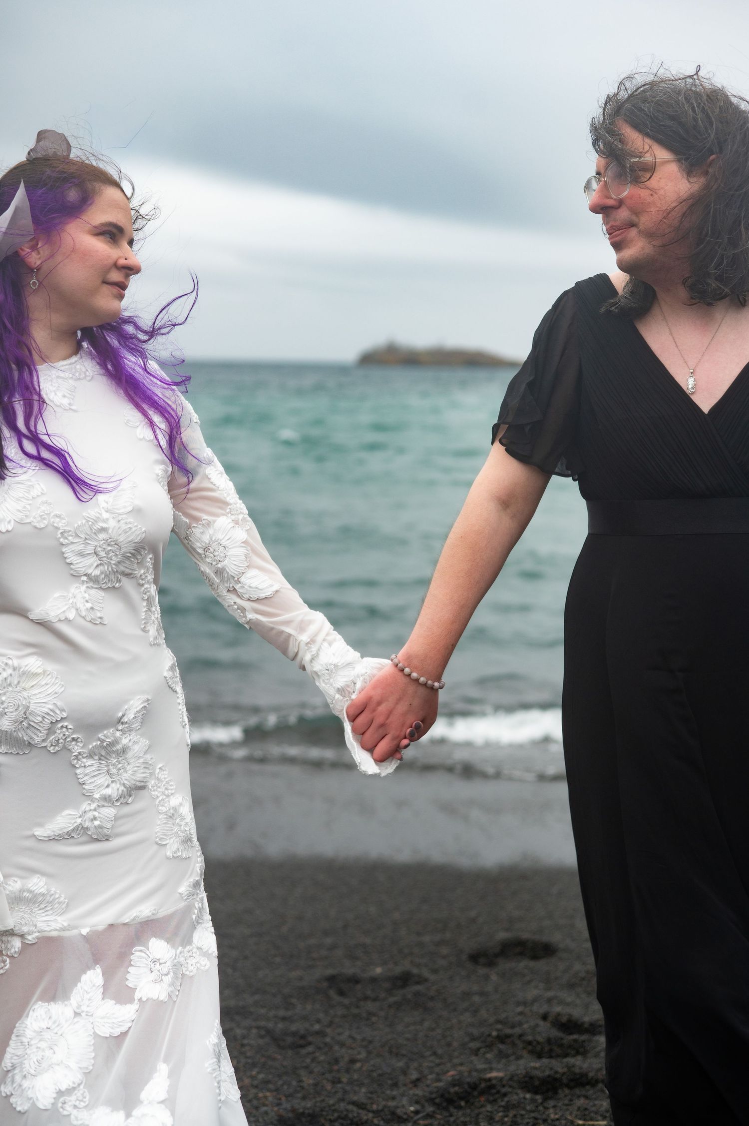 Two brides hold hands on a black sand beach, one in a white lace gown with purple hair, the other in a black dress.