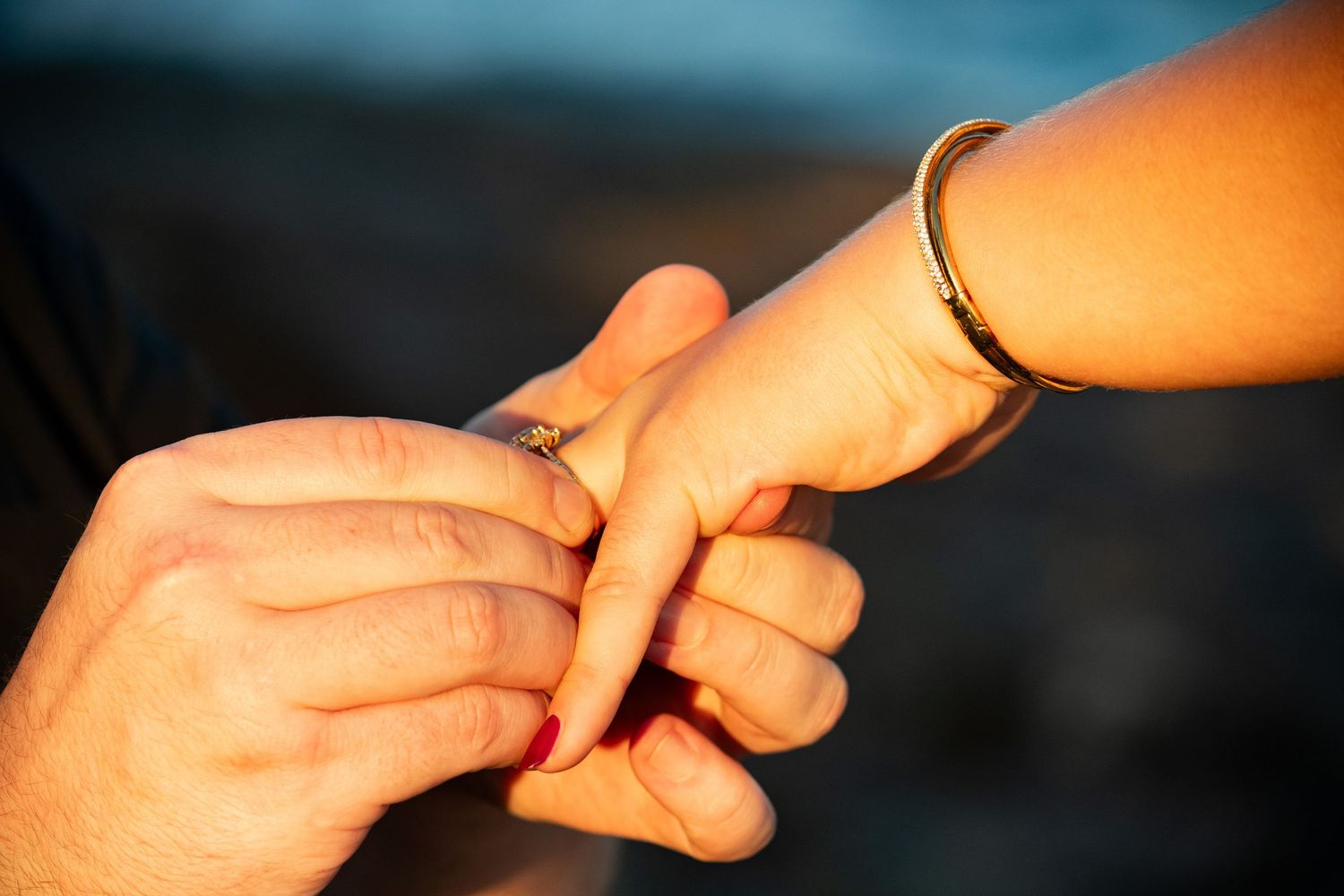Close-up of engagement ring being placed on woman's finger at golden hour by the water.