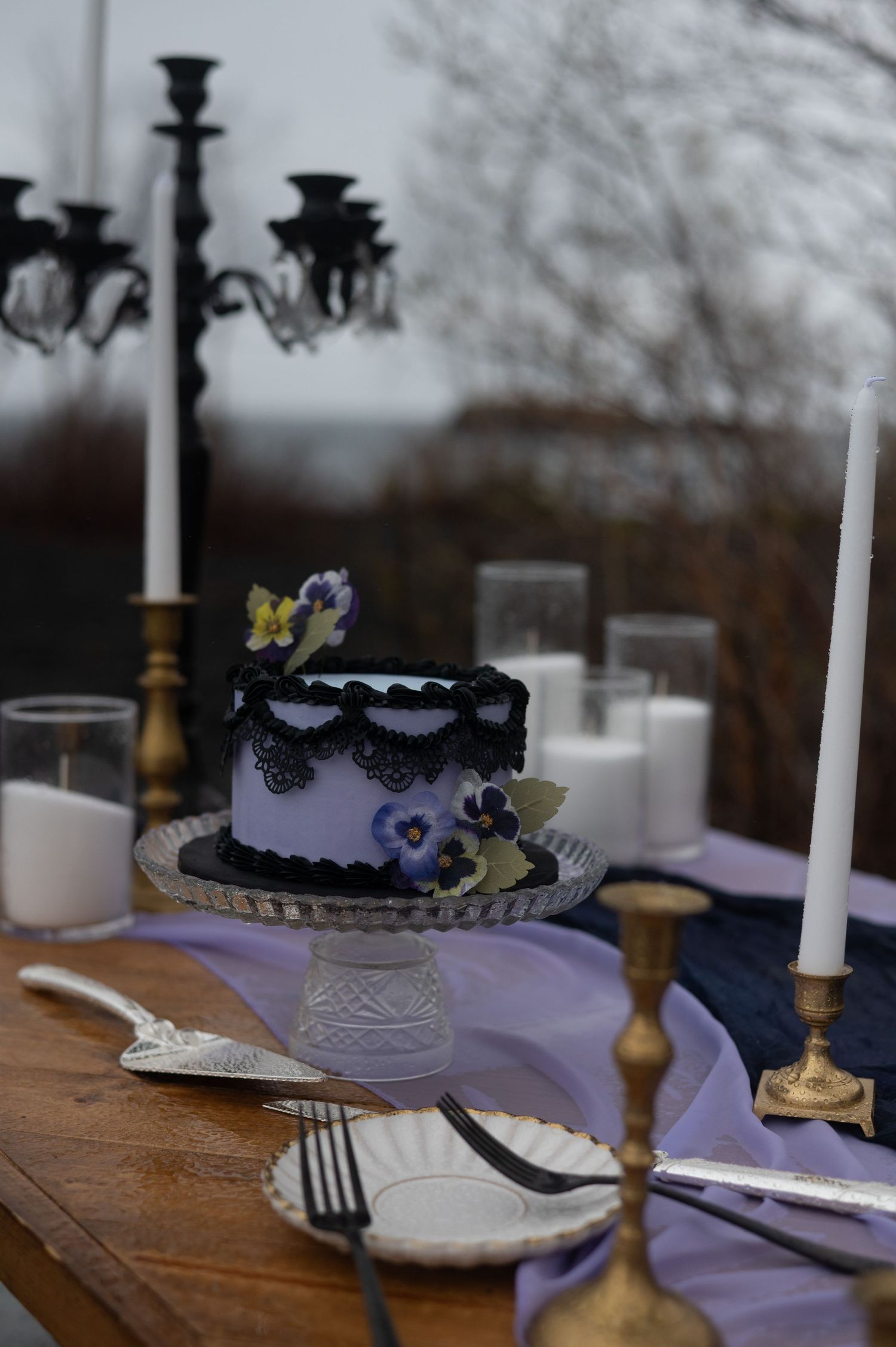 Elegant purple cake with floral decor on crystal stand, surrounded by gold candlesticks and white candles on rustic outdoor table.
