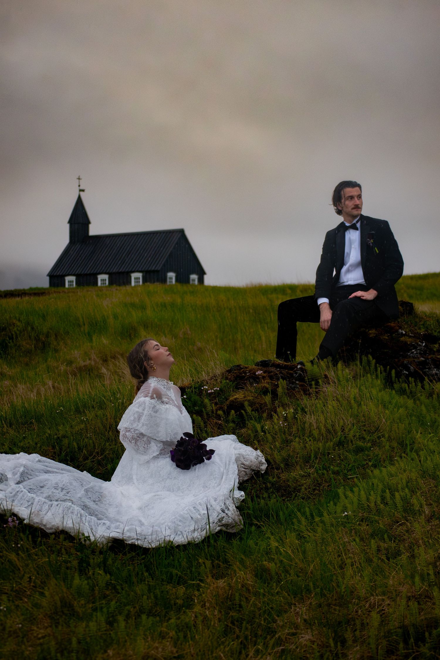 Bride in white lace gown sits on grass while groom in tuxedo perches on rocks near black Icelandic church.