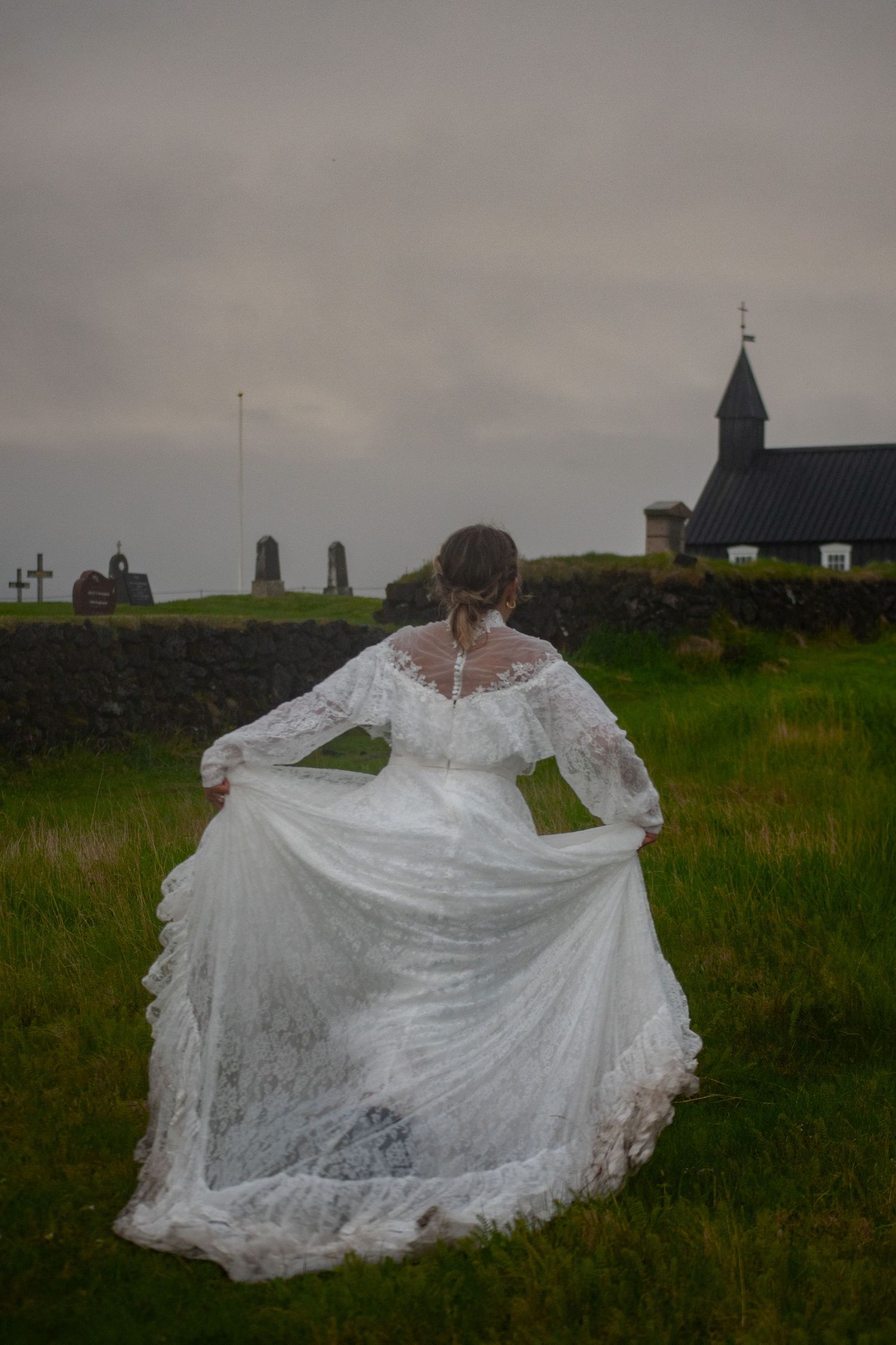 Bride in elegant lace wedding gown walking toward historic black church and cemetery on overcast day in Iceland.