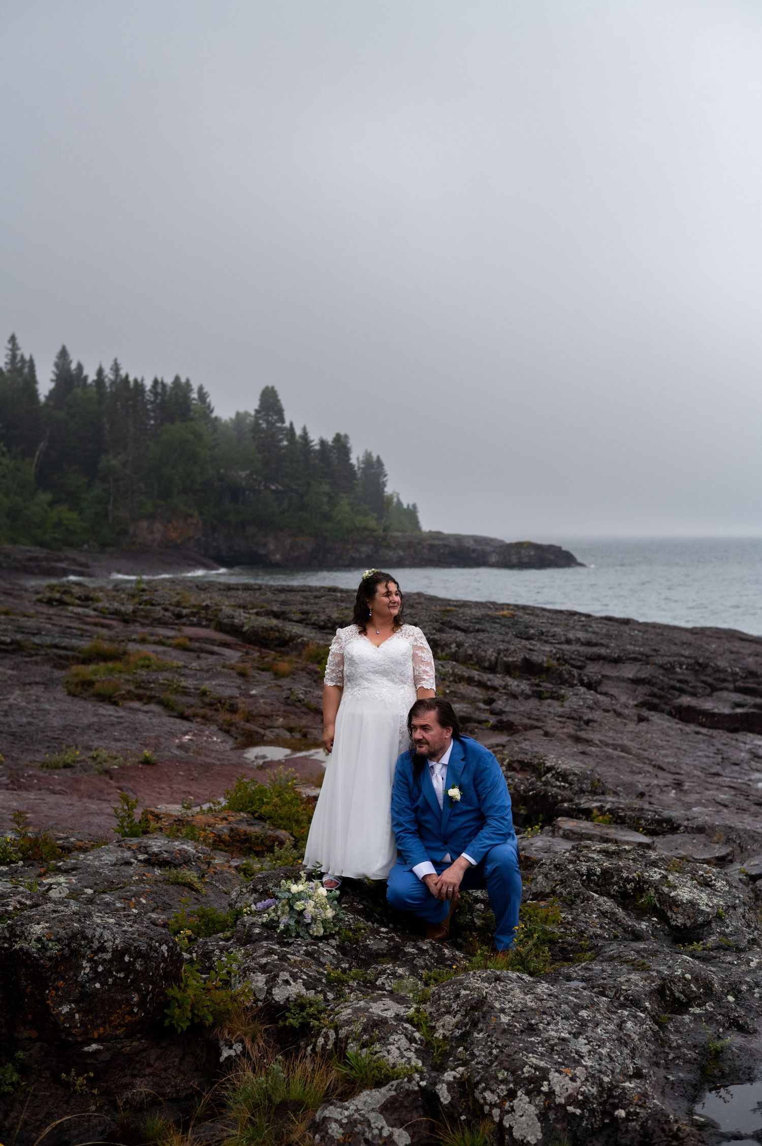 Bride in white lace gown and groom in blue suit pose on rocky coastal shoreline with evergreen trees and misty ocean backdrop.