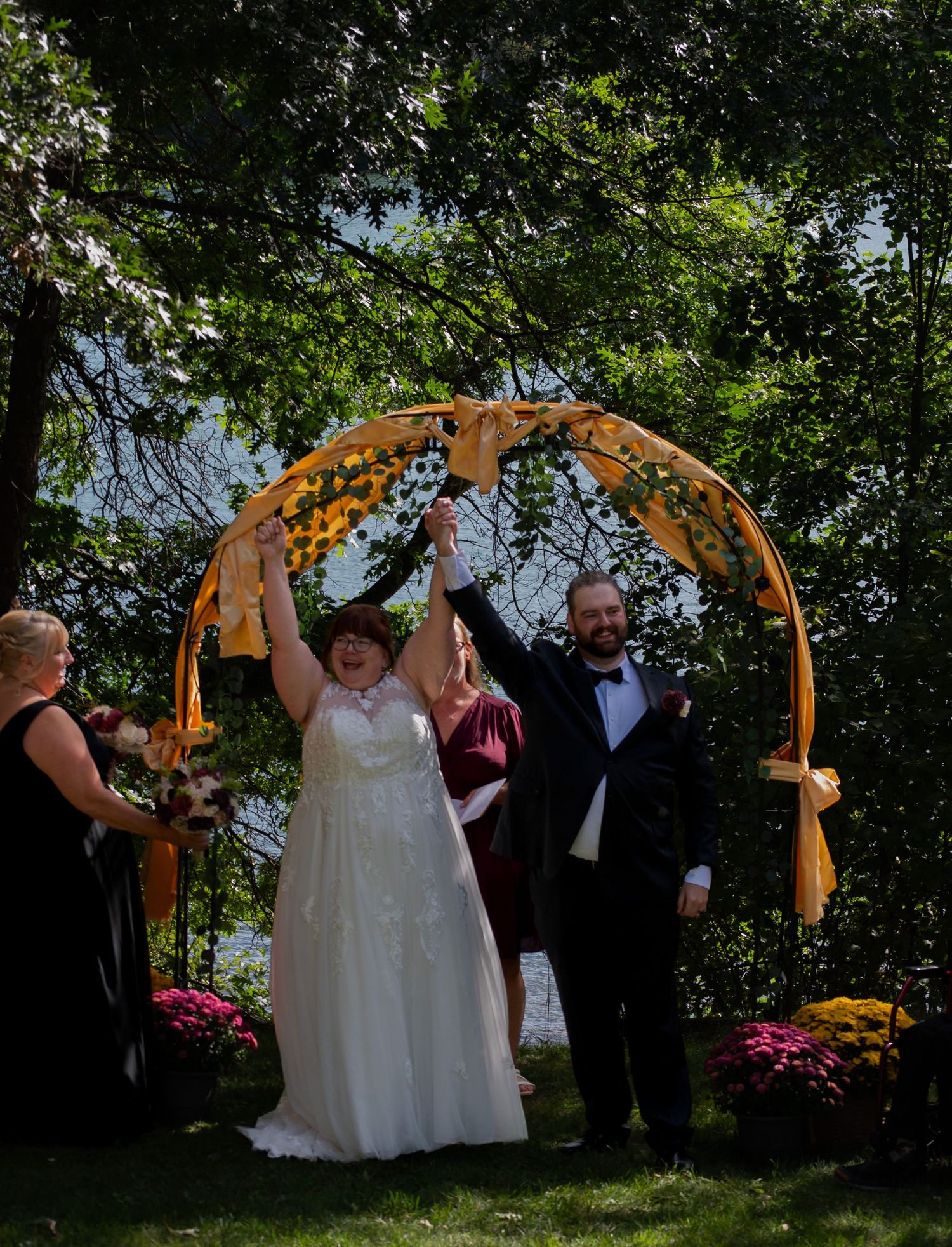 Joyful newlyweds raise hands under floral arch at outdoor wedding ceremony surrounded by lush greenery and colorful flowers.