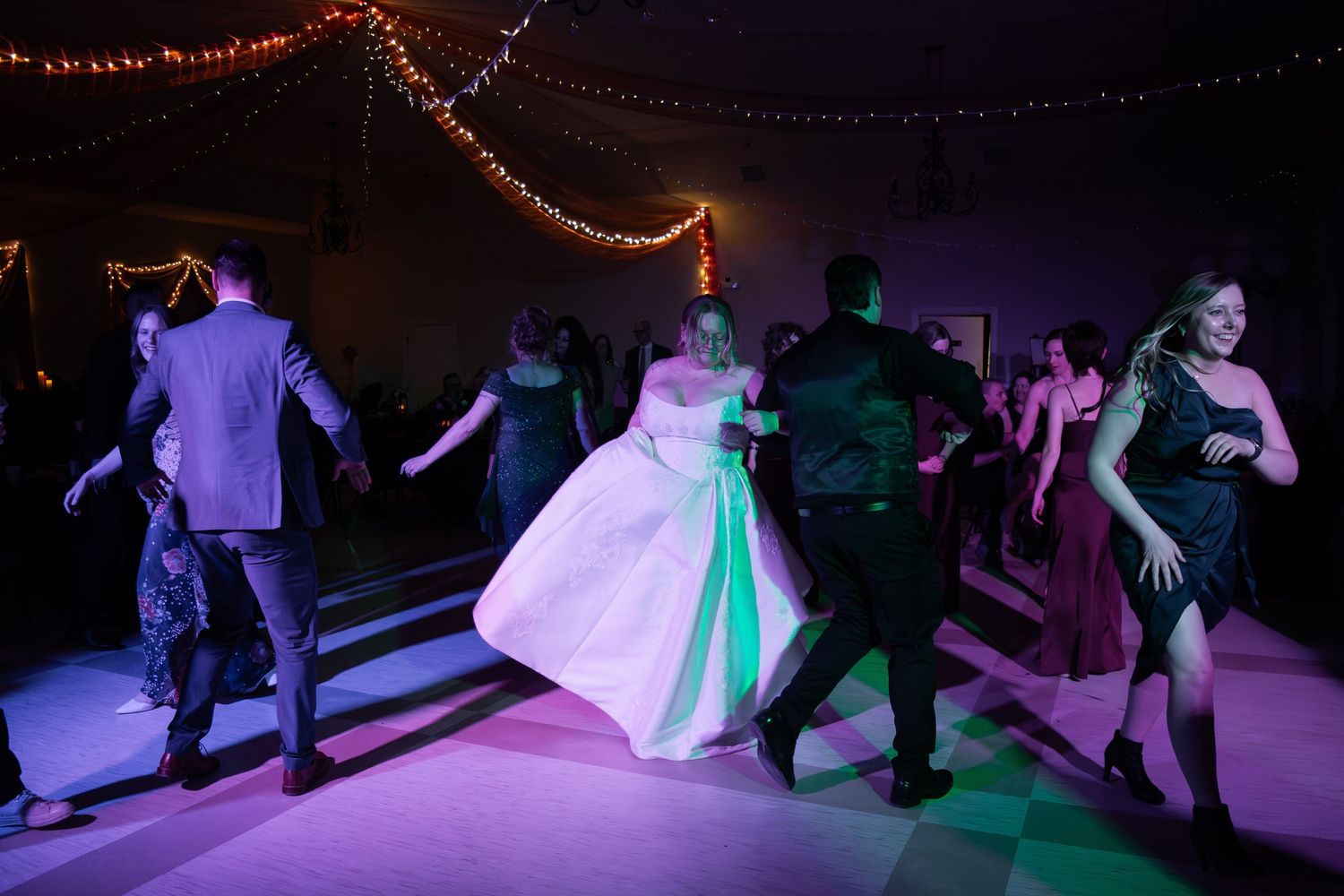 Bride and wedding guests dancing together on a decorated dance floor with purple and green lighting.