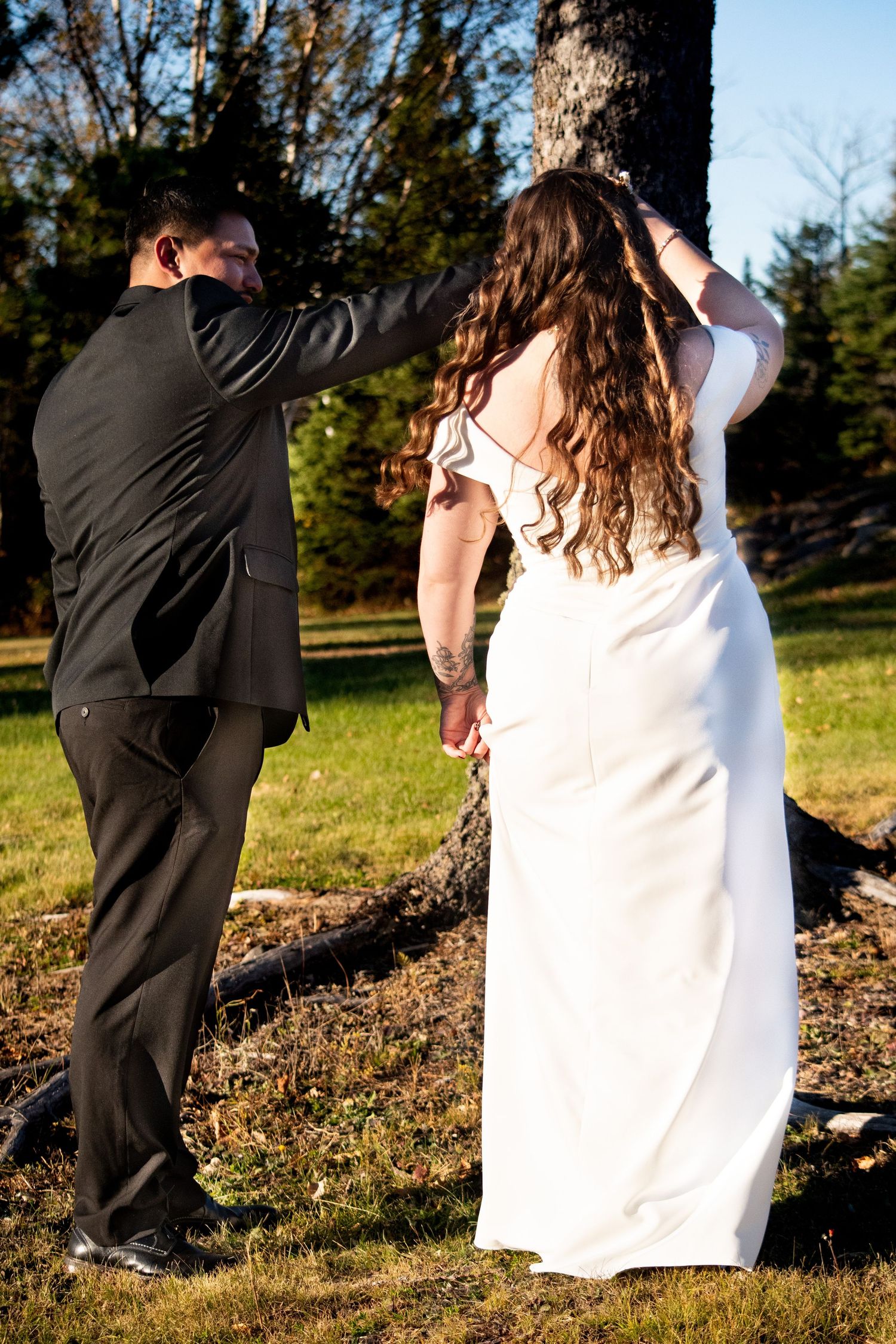 Bride in white backless gown and groom in black suit holding hands walking toward tree outdoors.