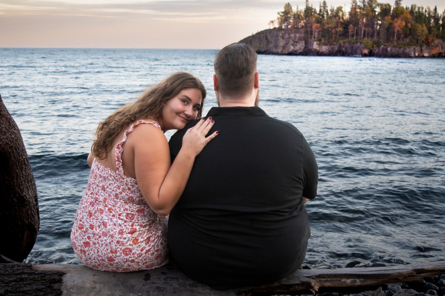 Couple sitting together on a log by the water, woman leaning affectionately on her partner.