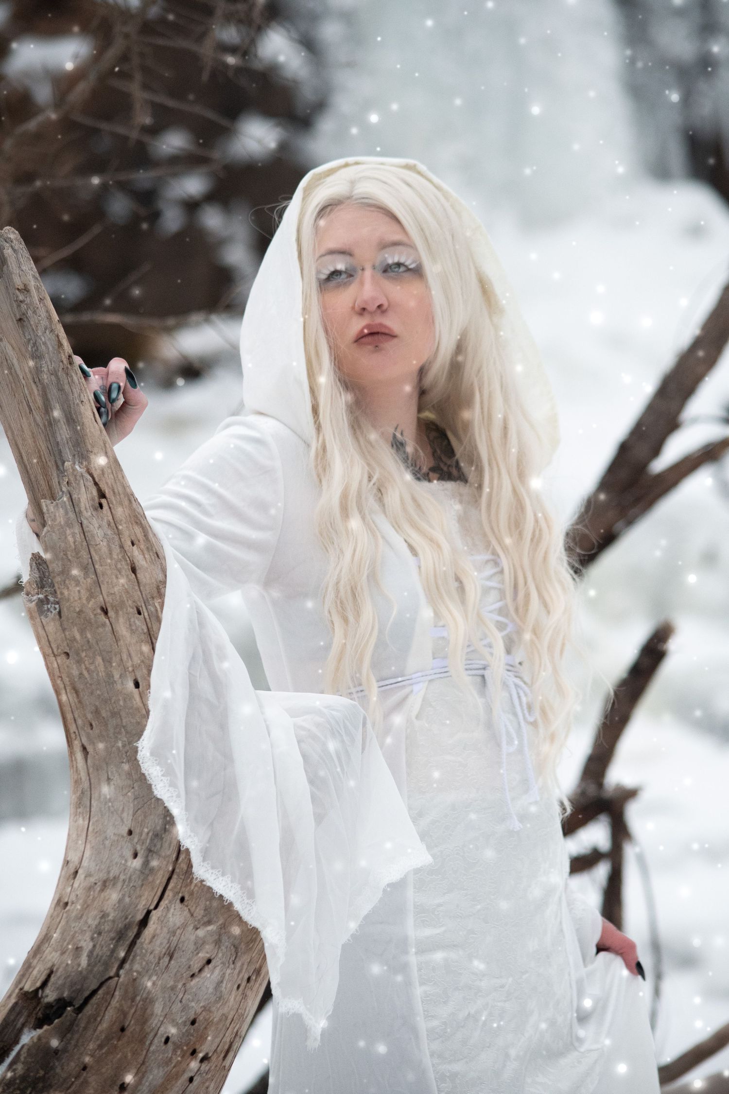 Tattooed blonde woman in white medieval gown posing by driftwood in snowy winter scene.