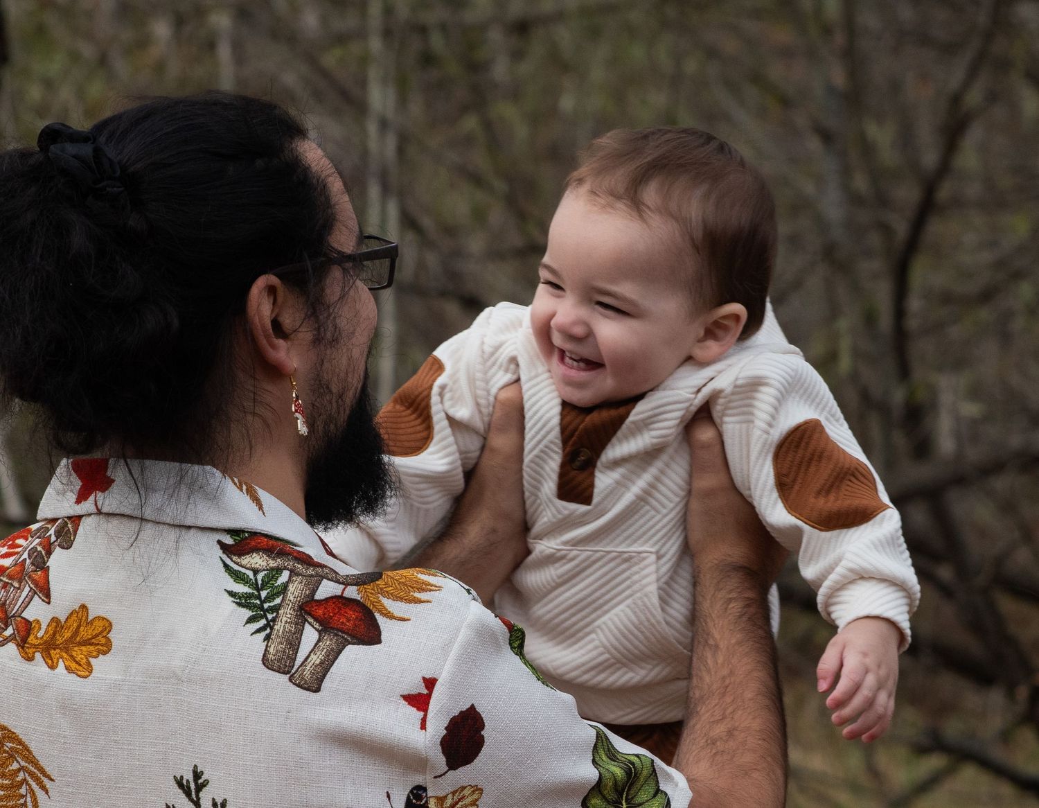 Father in floral shirt lifting giggling baby face-to-face outdoors in nature.