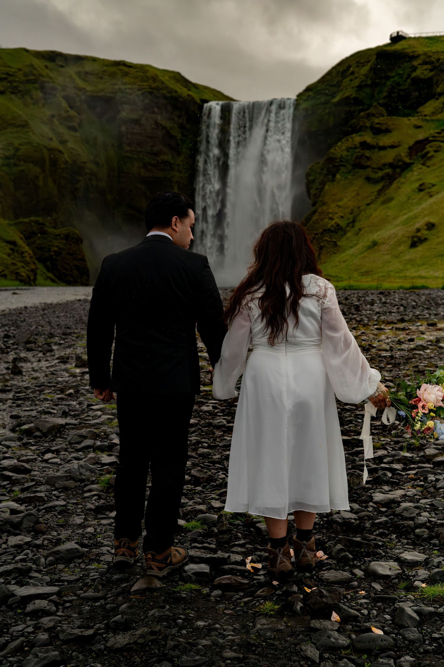 Couple holding hands facing Skogafoss waterfall in Iceland, bride in white dress with bouquet, groom in black suit.