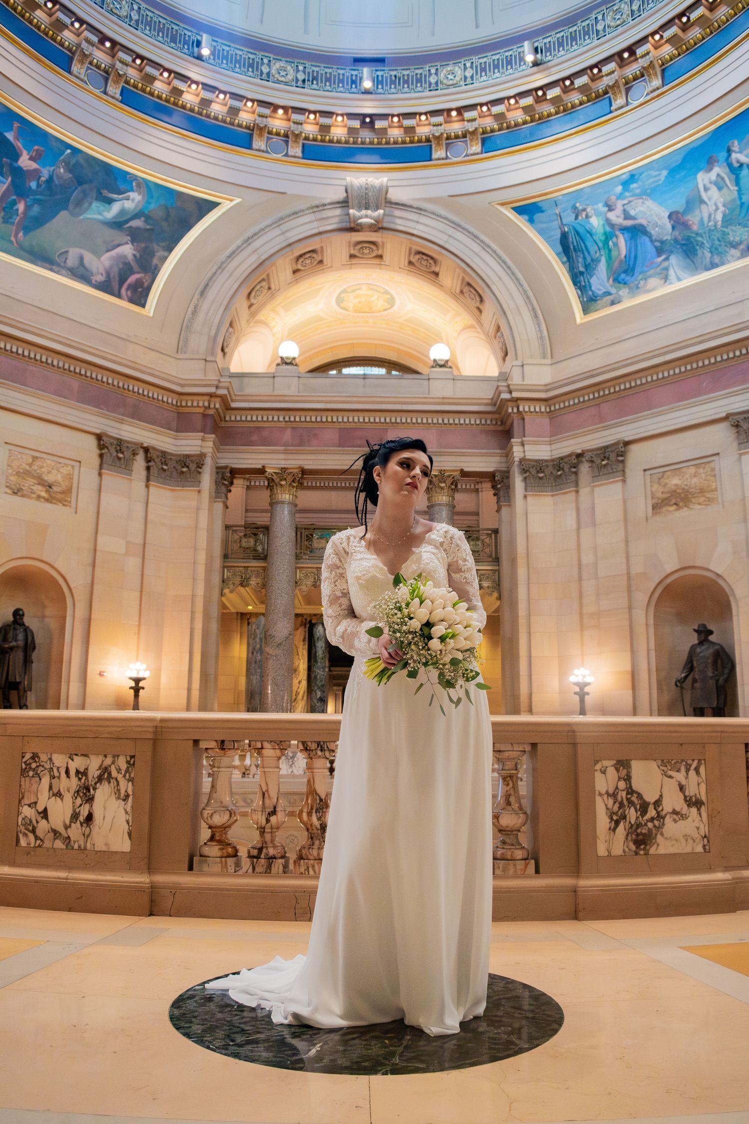 Bride in lace wedding gown holds bouquet beneath stunning painted rotunda dome of a grand capitol building.