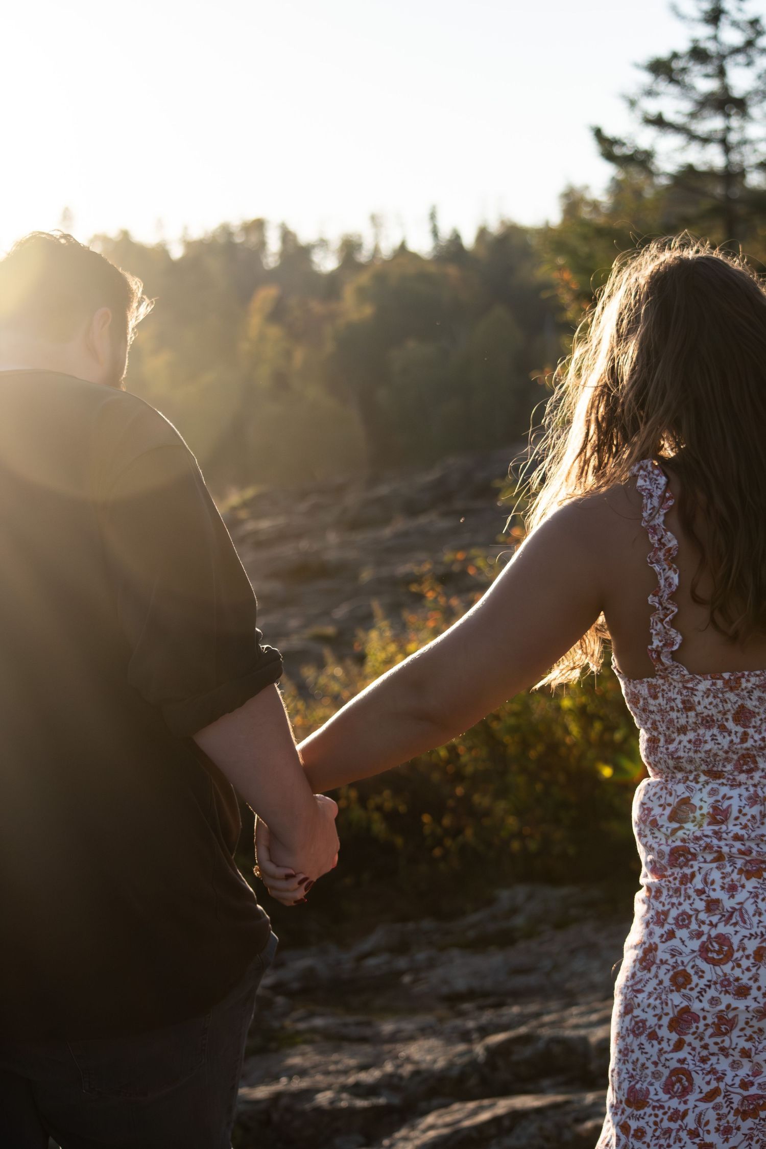 Couple holds hands walking toward sunset on rocky landscape surrounded by trees.