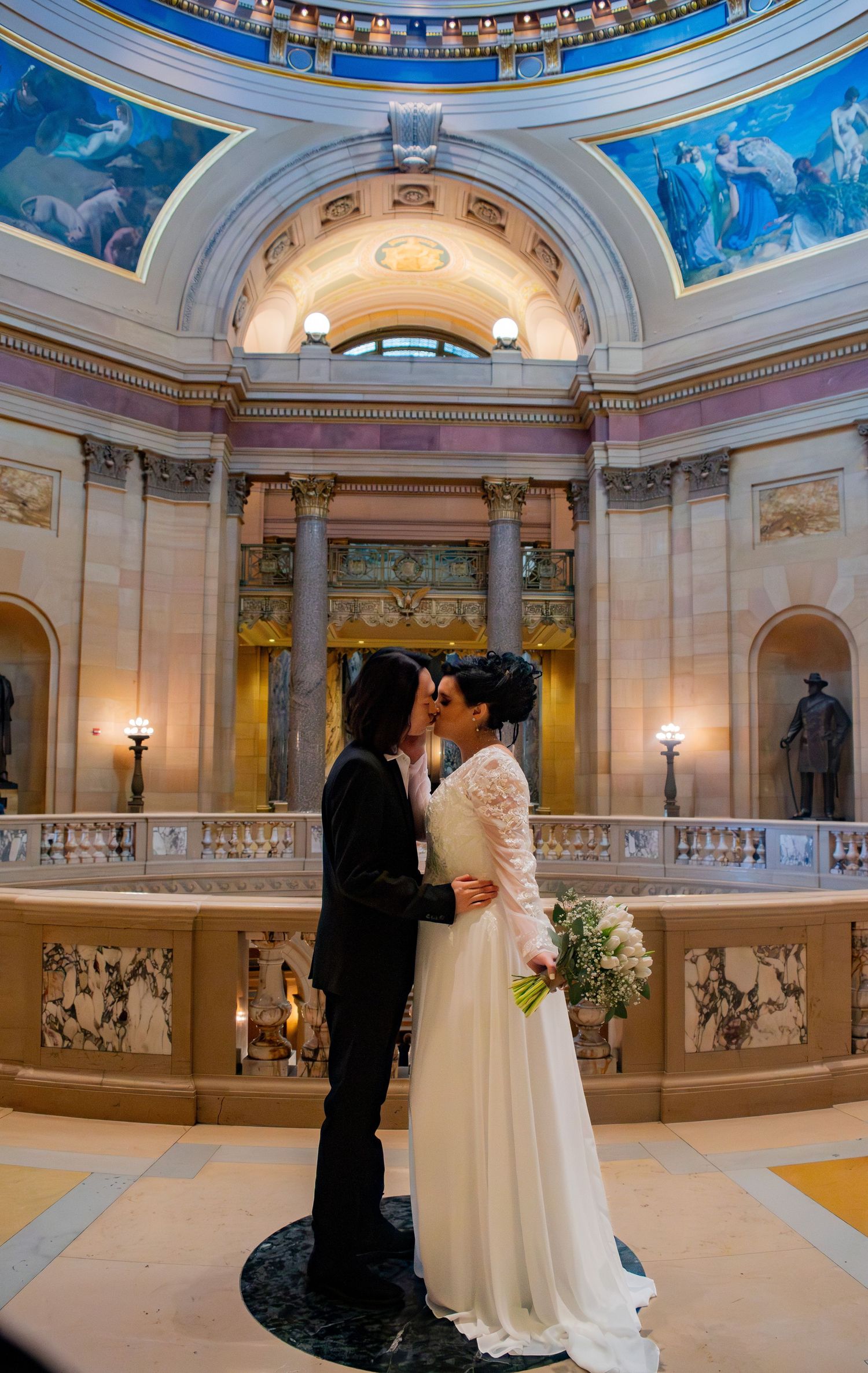 Same-sex couple embraces romantically inside grand rotunda with painted murals and classical architecture.