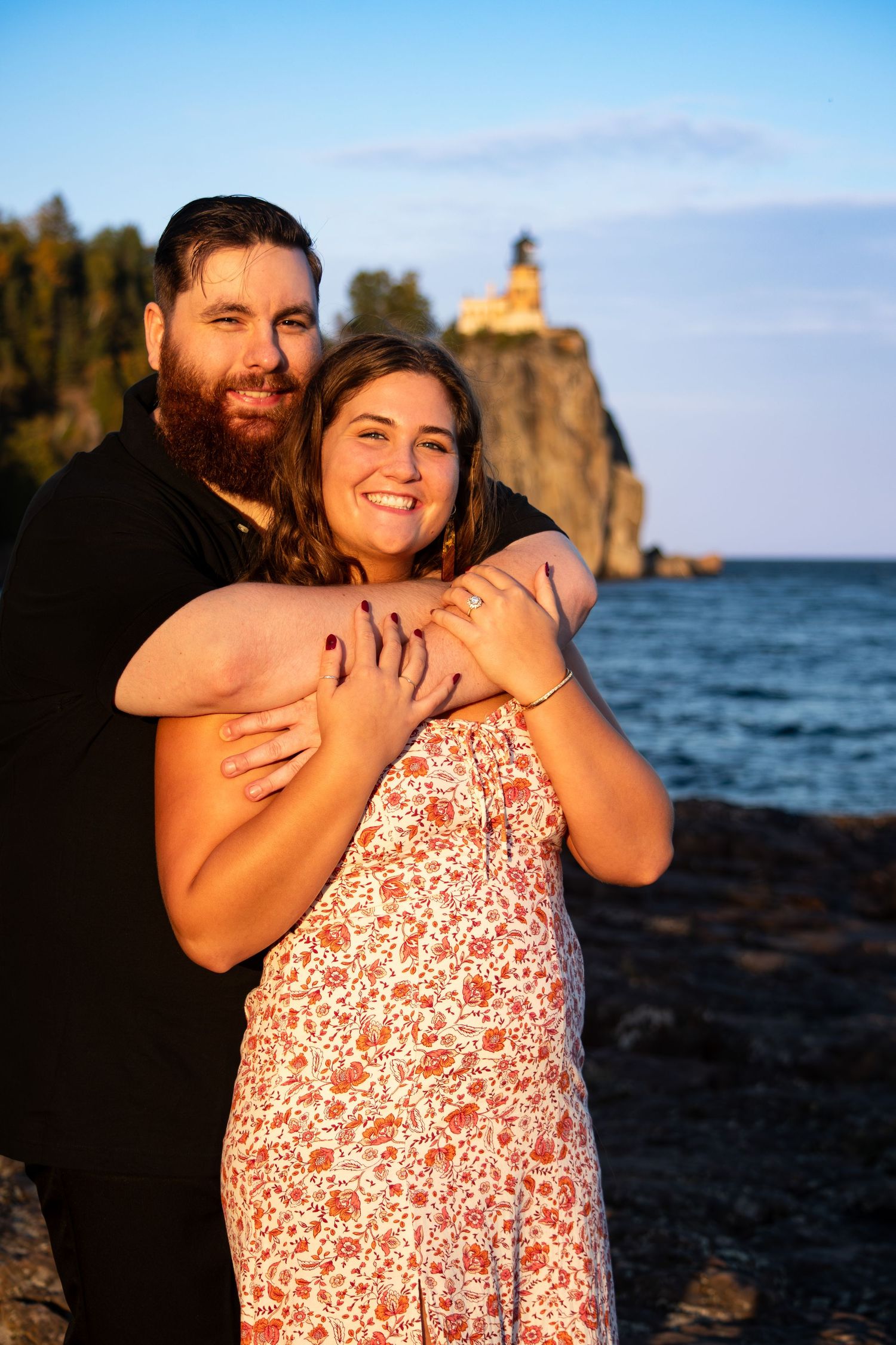 Smiling engaged couple embraces at golden hour with Split Rock Lighthouse in background.