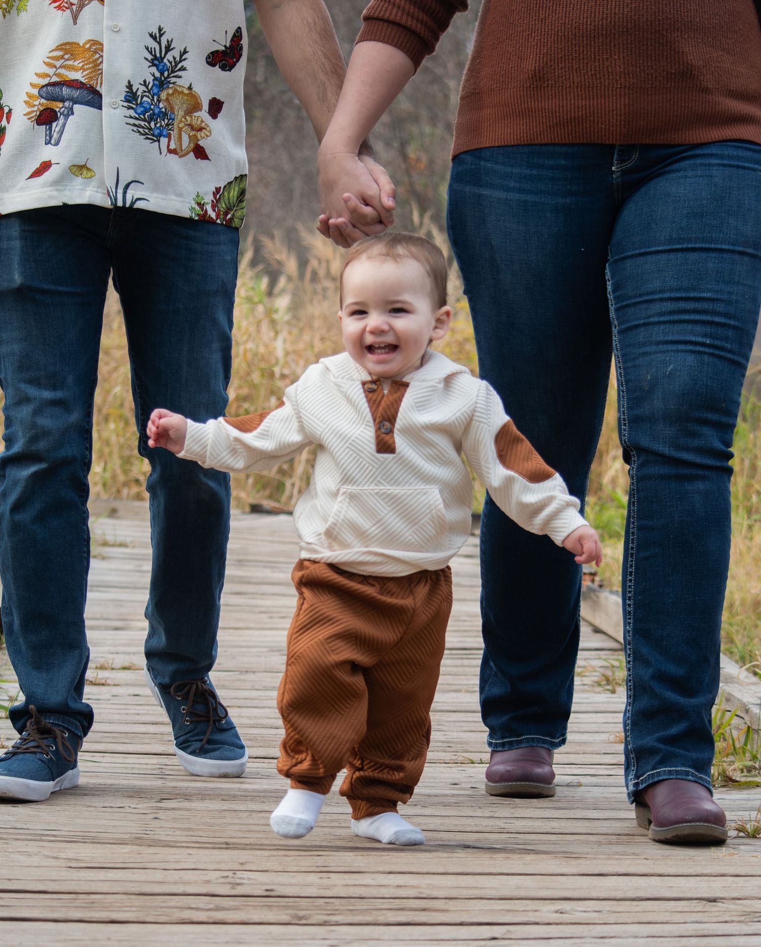 Happy toddler boy in cream sweater and brown pants walking on wooden boardwalk held by parents.