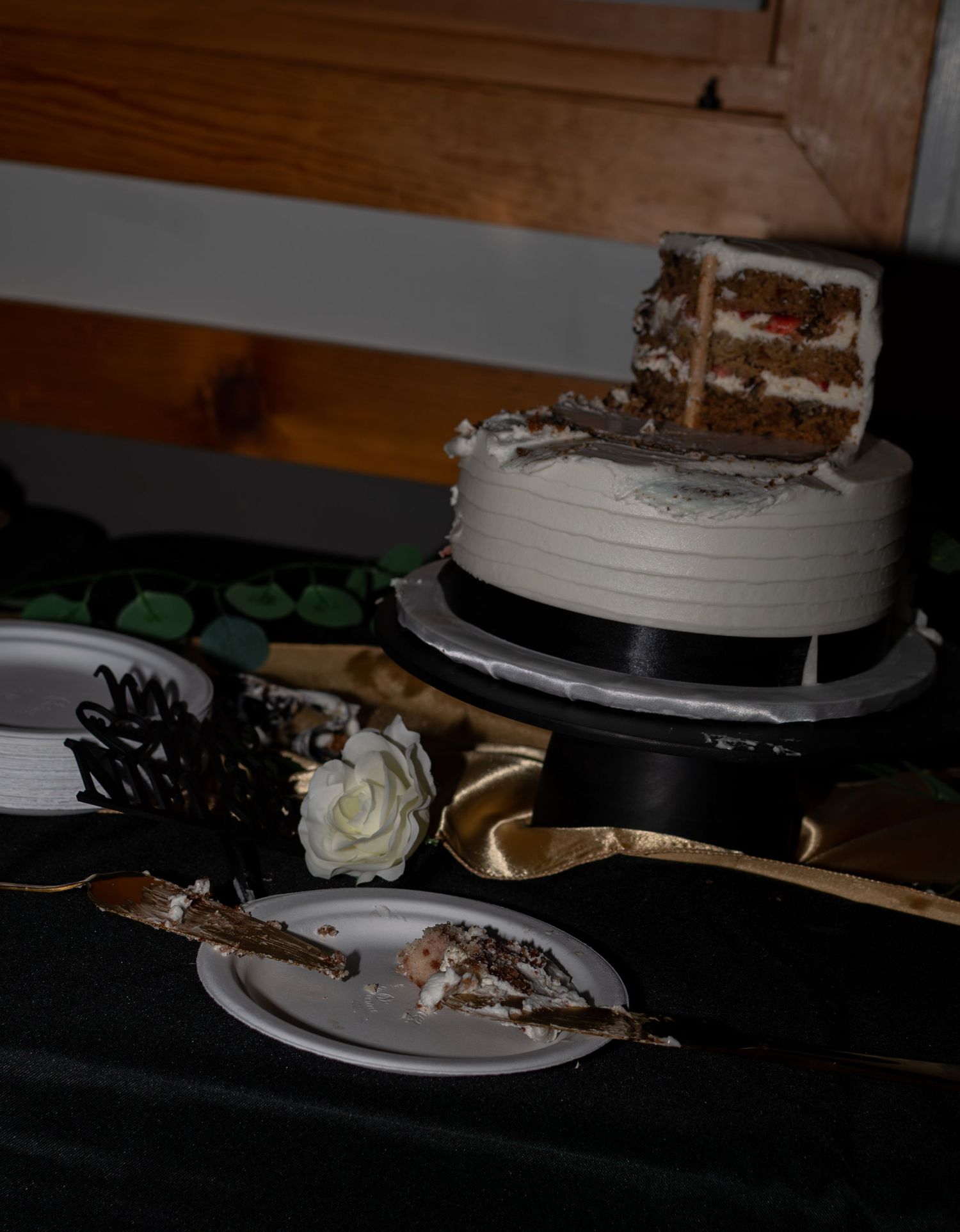 Partially sliced white frosted layer cake on a black stand with a used cake server and white rose on a dark table.
