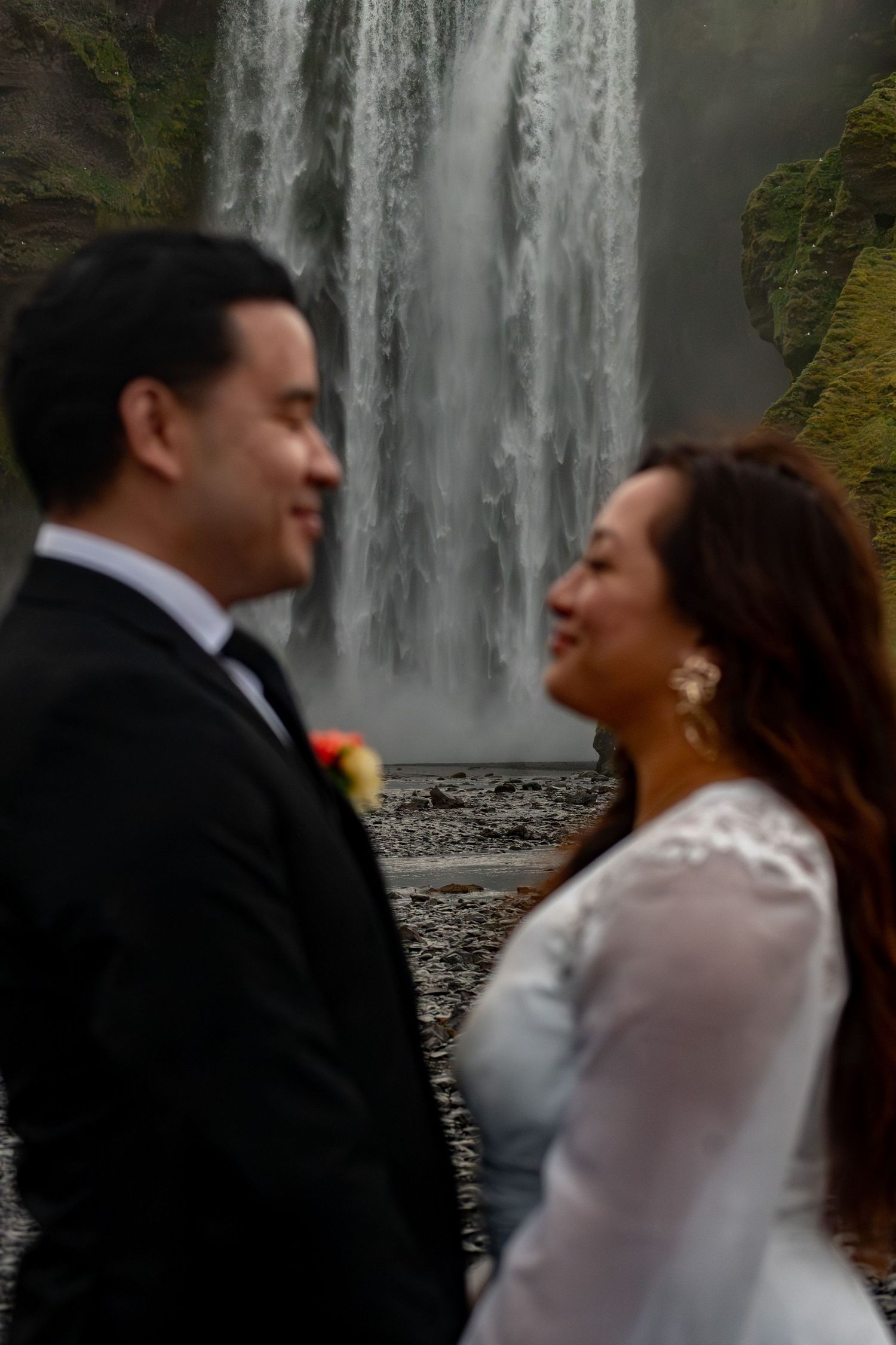 A couple in wedding attire smiles at each other in front of a stunning waterfall backdrop.