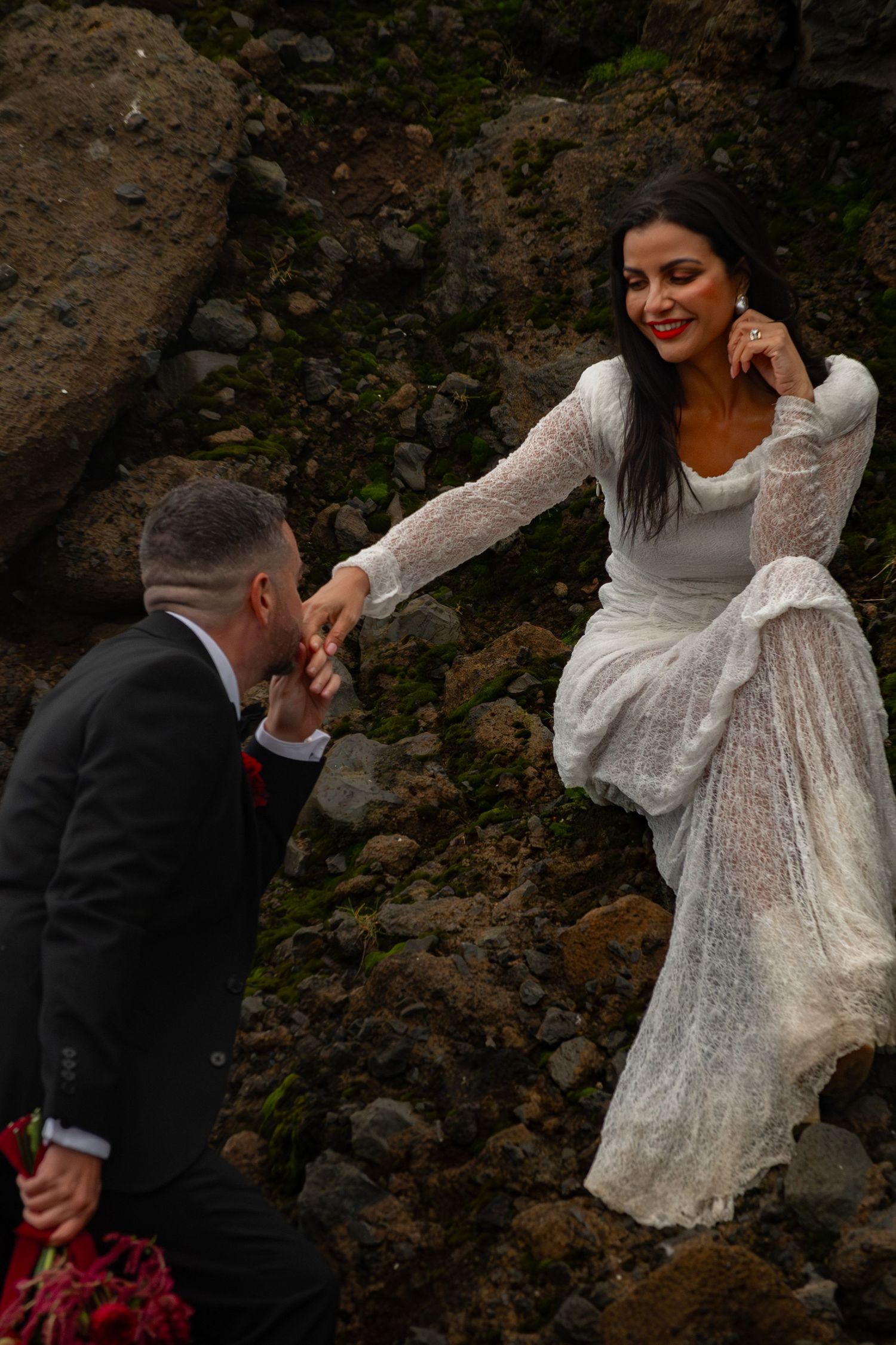 A bride in a white lace gown holds hands with a suited groom among rocky terrain, smiling joyfully.