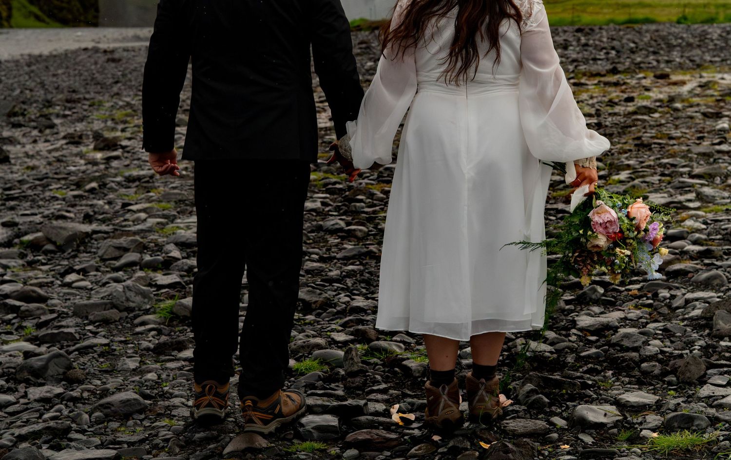 Bride and groom holding hands on rocky beach, woman in white dress holding bouquet, viewed from behind.