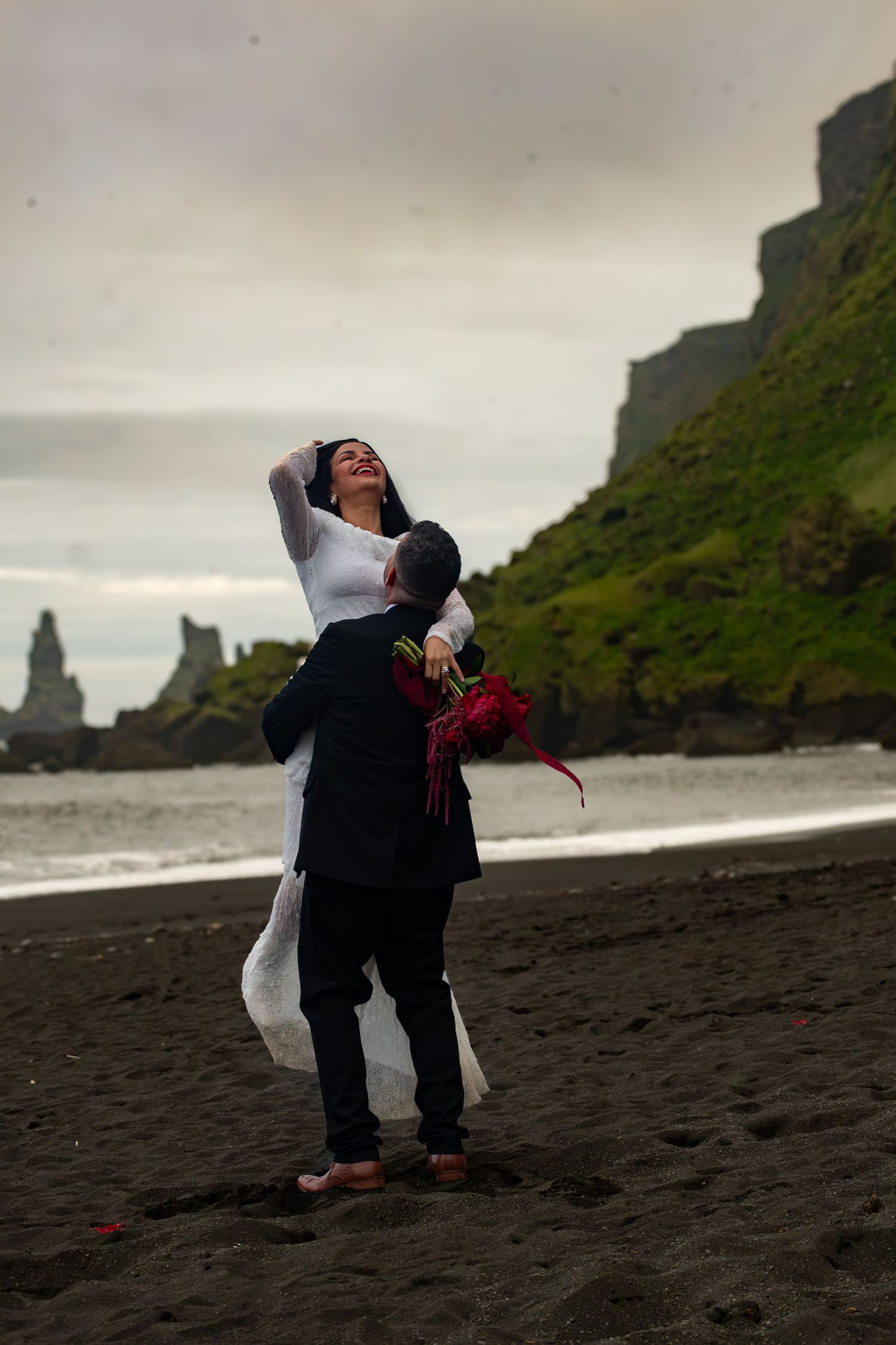 Couple in wedding attire embracing on black sand beach with dramatic cliffs and ocean backdrop, bride holding red roses.