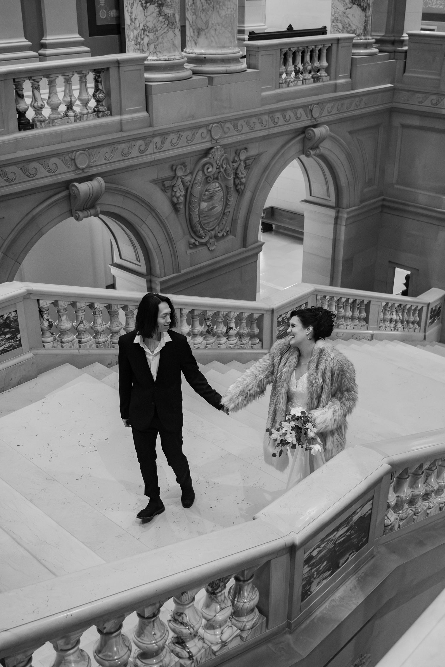 Black and white photo of couple holding hands on grand ornate marble staircase in historic building.