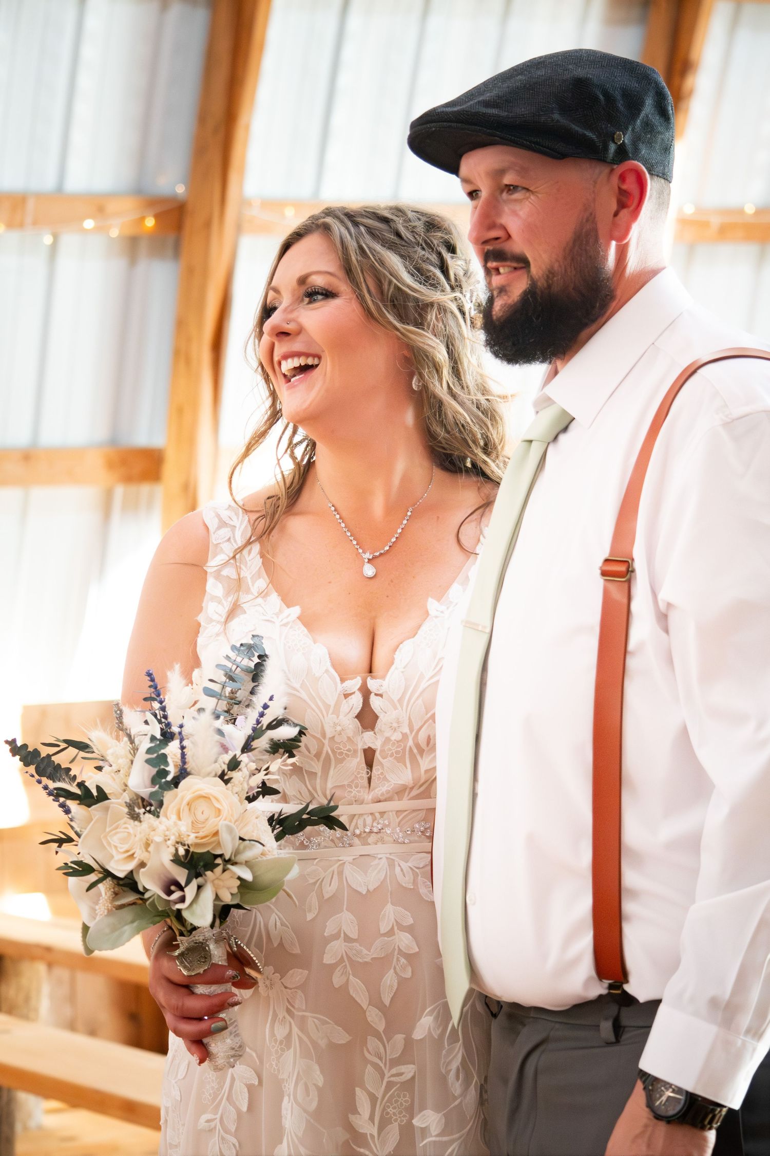 Smiling bride holding white floral bouquet standing beside bearded groom with suspenders in a barn venue.