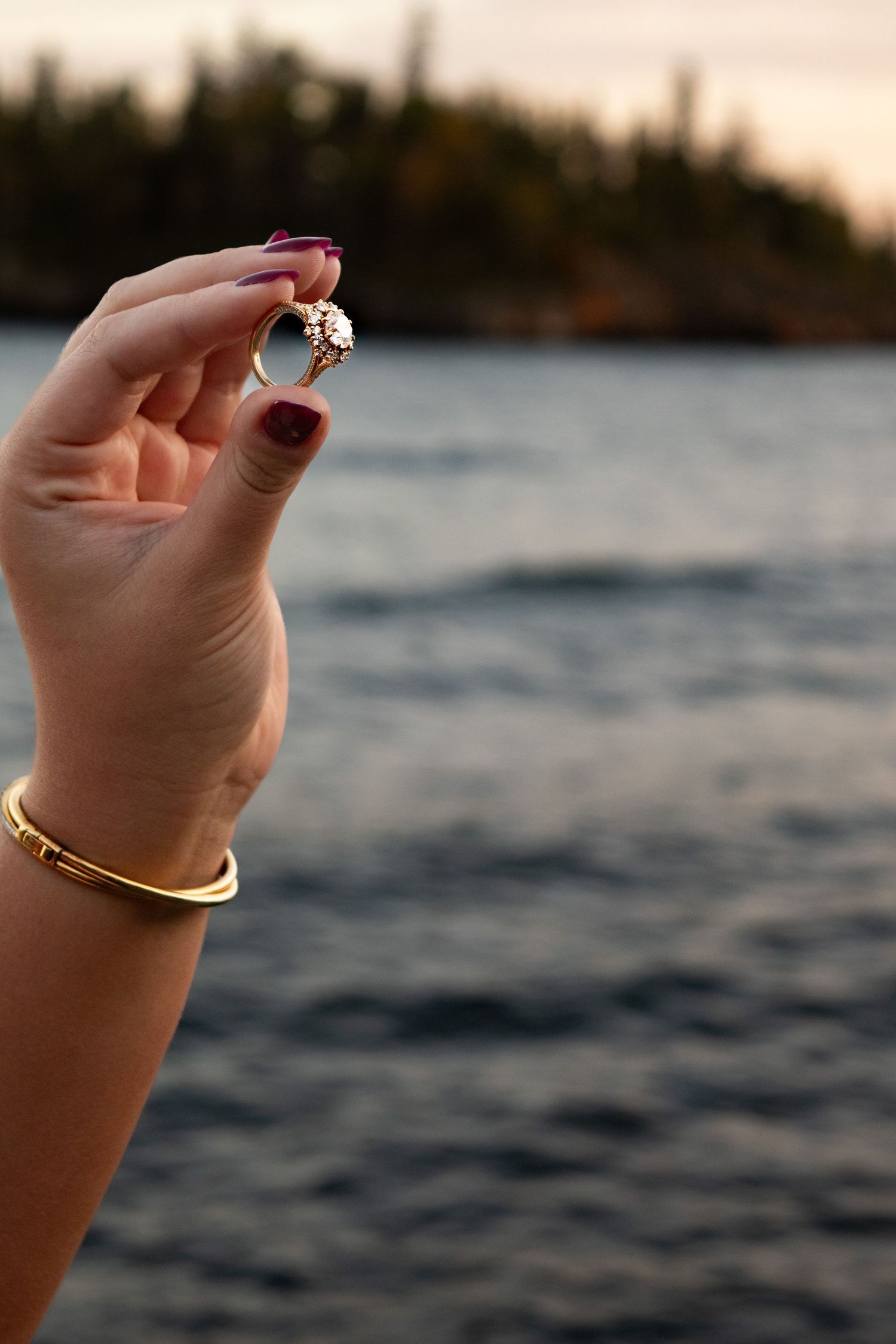 Woman holds up diamond engagement ring near a lake at sunset with trees in background.