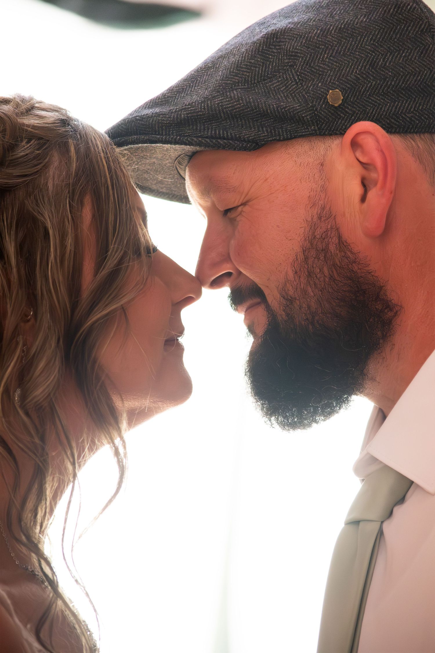 Romantic close-up of bearded groom in flat cap and bride touching noses in tender wedding moment.