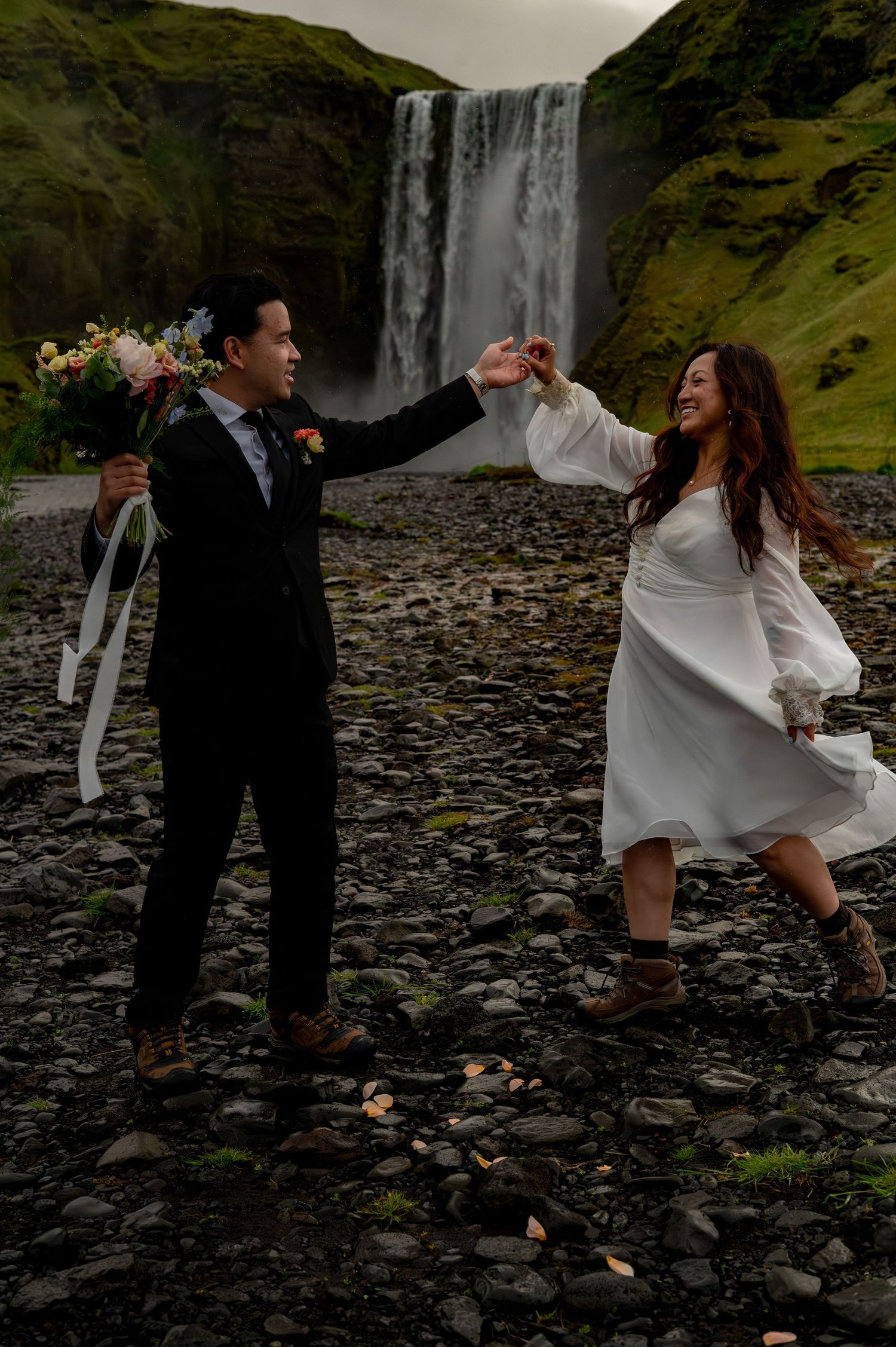 Couple eloping at Skogafoss waterfall in Iceland, groom in suit holding bouquet while bride in white dress twirls on rocky shore.
