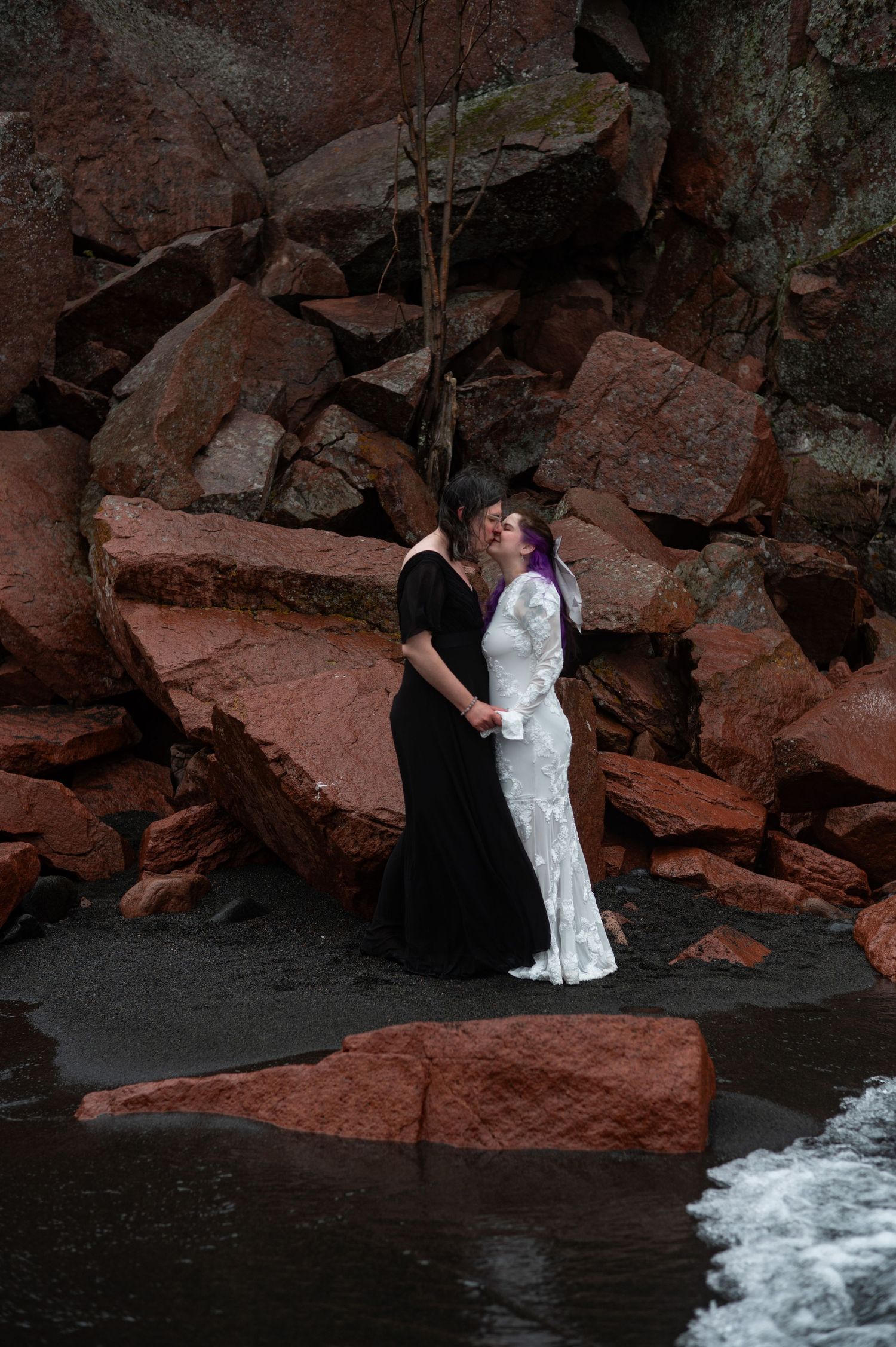 A couple in wedding attire kisses romantically among red rocks on a dark beach, bride in white lace gown with purple hair.