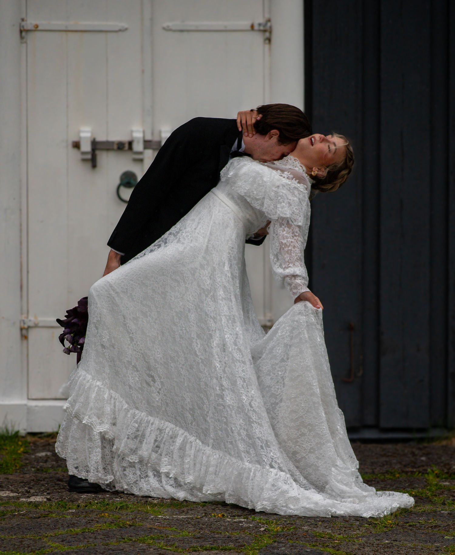 A groom in a black tuxedo dips and kisses his bride in a white lace wedding dress outdoors.