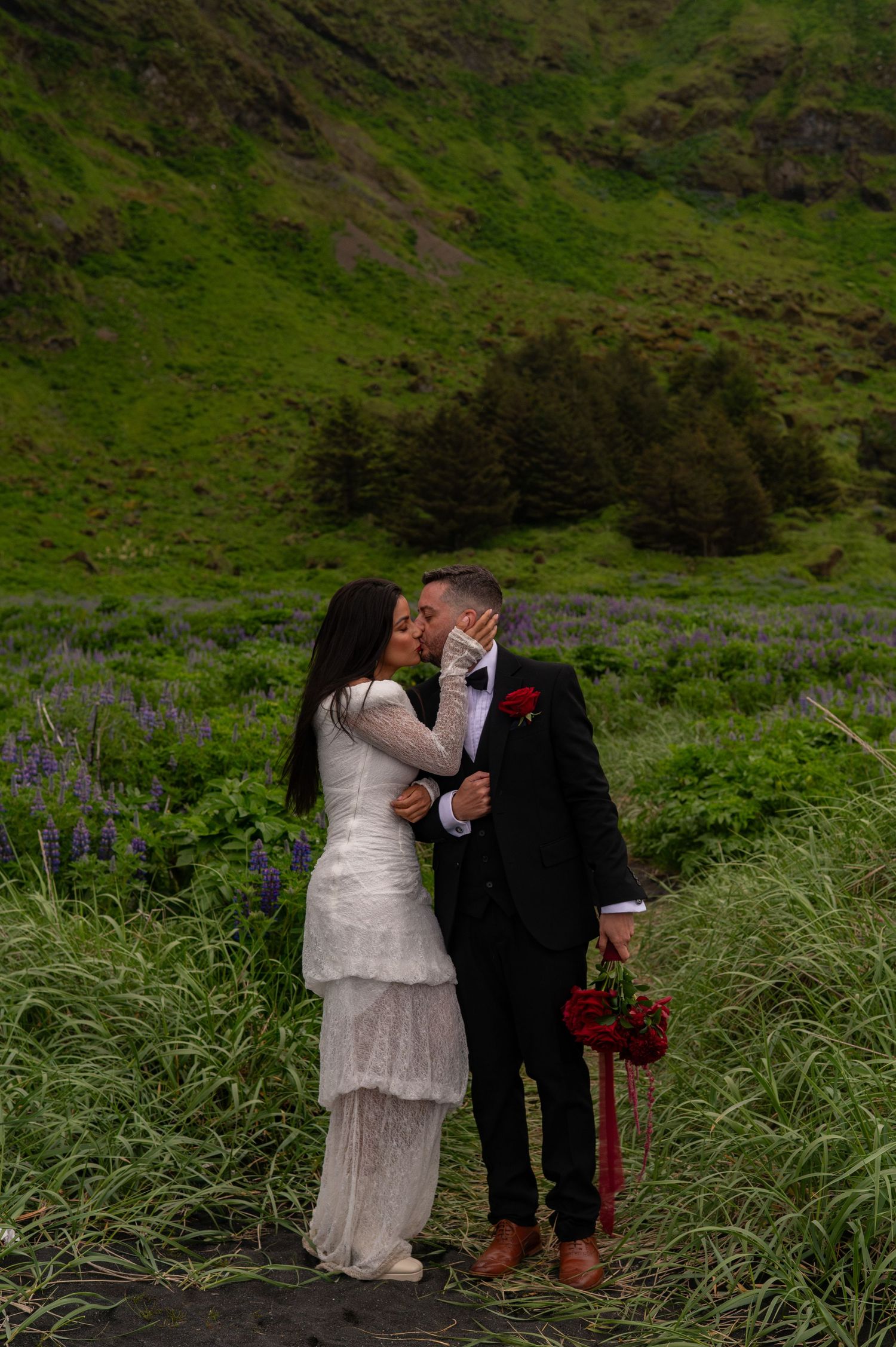 A couple shares a kiss in a lush green field with purple wildflowers, bride in white dress, groom in black suit.