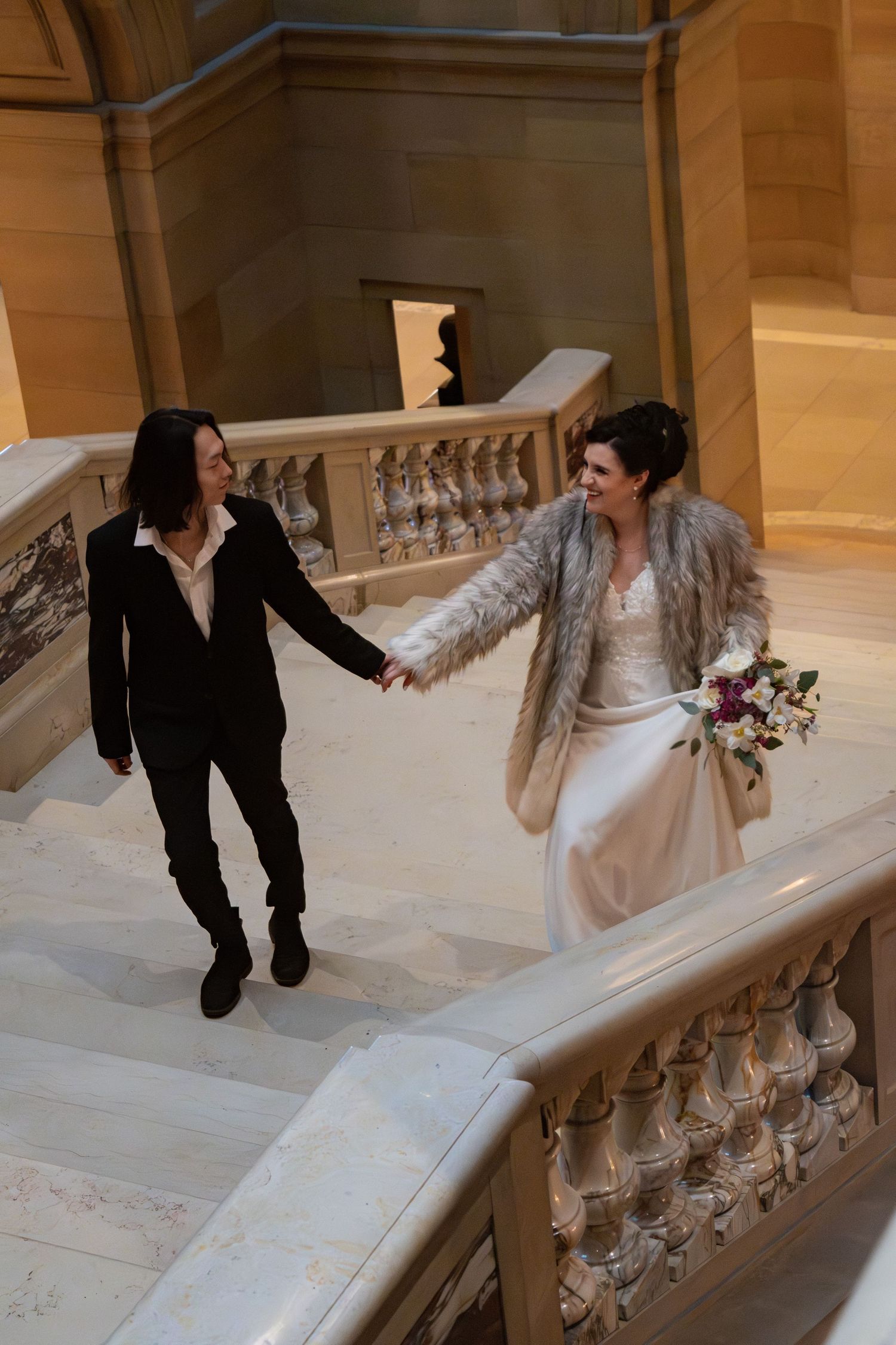 Two brides hold hands walking up marble stairs in an ornate building, one in a white gown with fur coat.