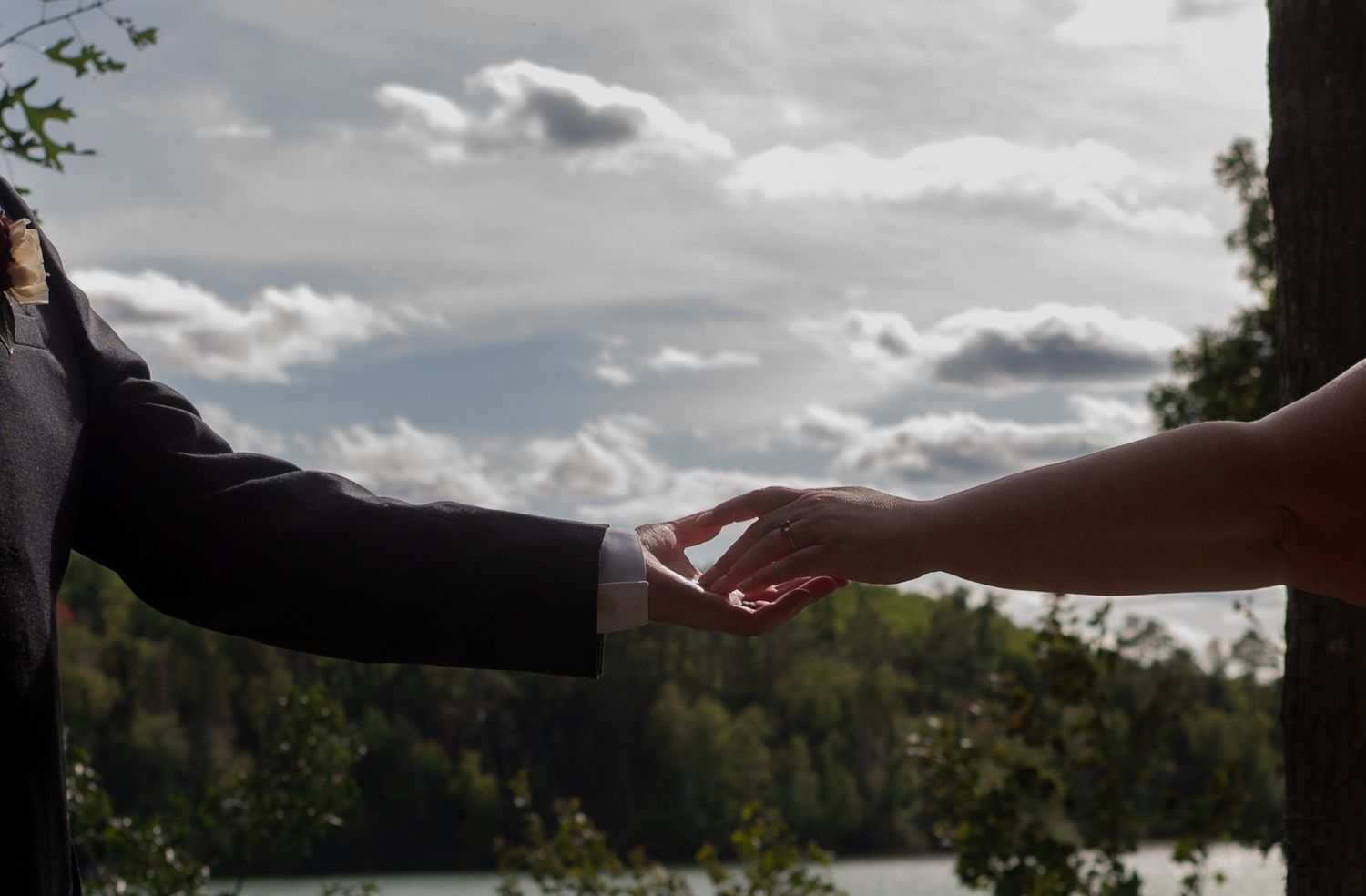 Two people reaching toward each other outdoors near a lake, with trees and cloudy sky in background.