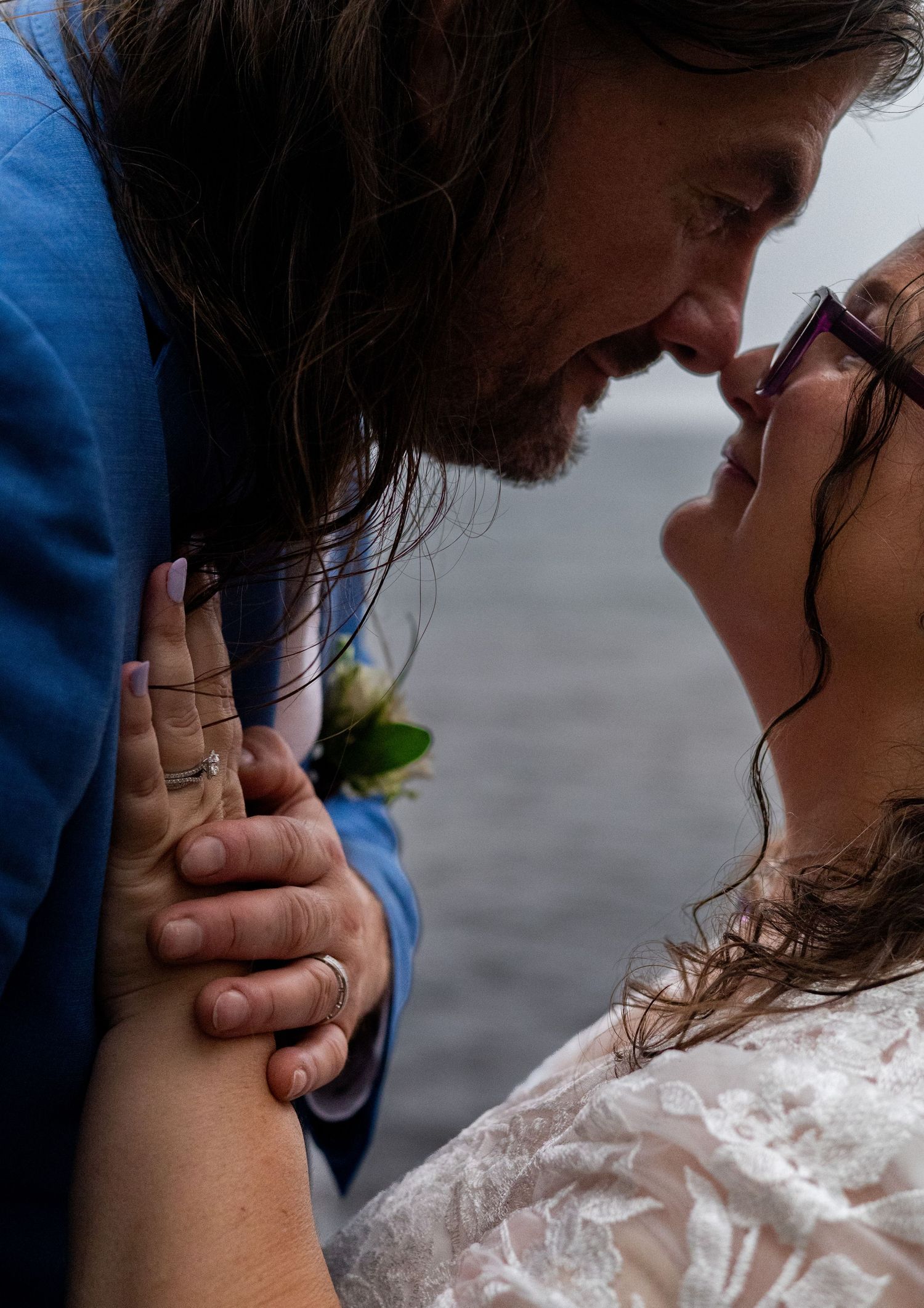 A bride and groom share a tender almost-kiss moment, hands intertwined, dressed in wedding attire near water.