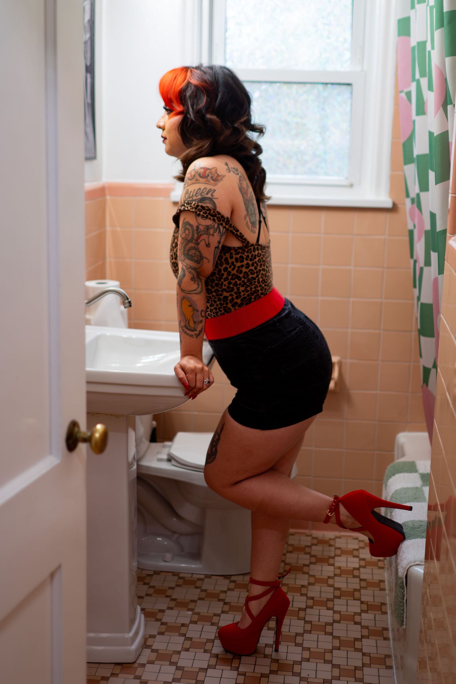 Tattooed woman in black mini skirt, leopard top, and red heels posing in a vintage pink tiled bathroom.
