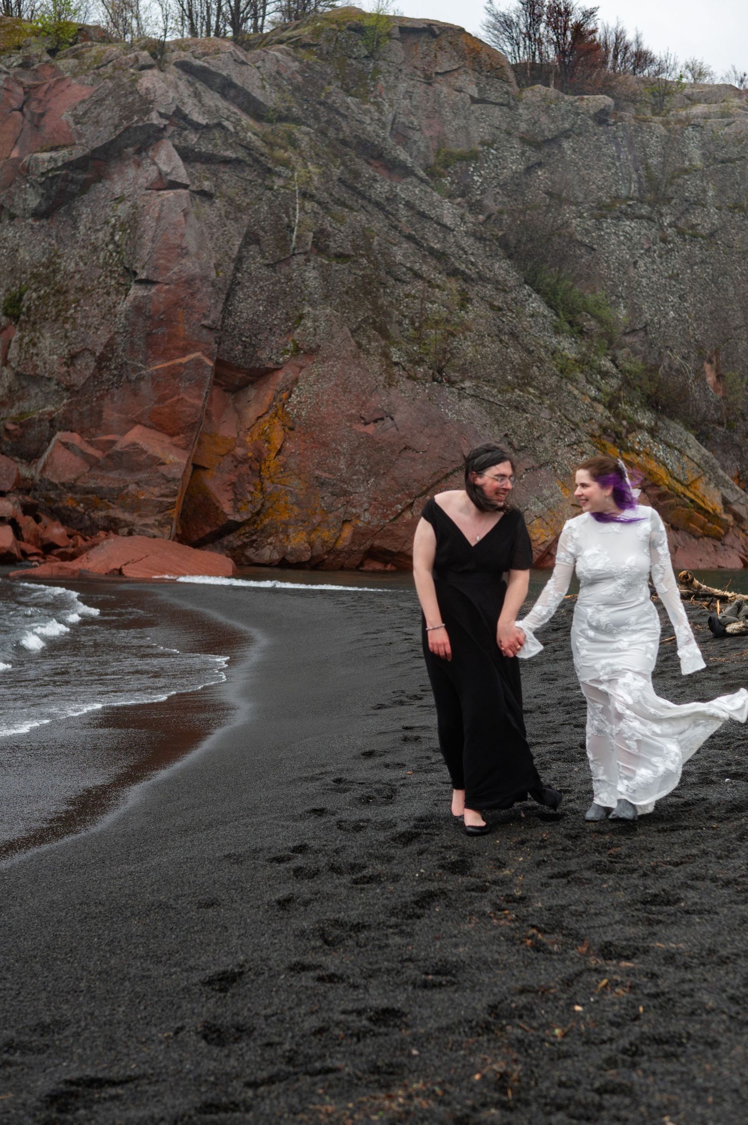 Two brides walk hand-in-hand on a black sand beach, one in white lace and one in black, with red rock cliffs behind.