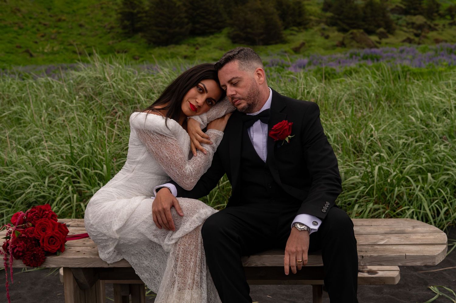 Bride in white lace gown and groom in black tuxedo sitting on bench, leaning together in green meadow.