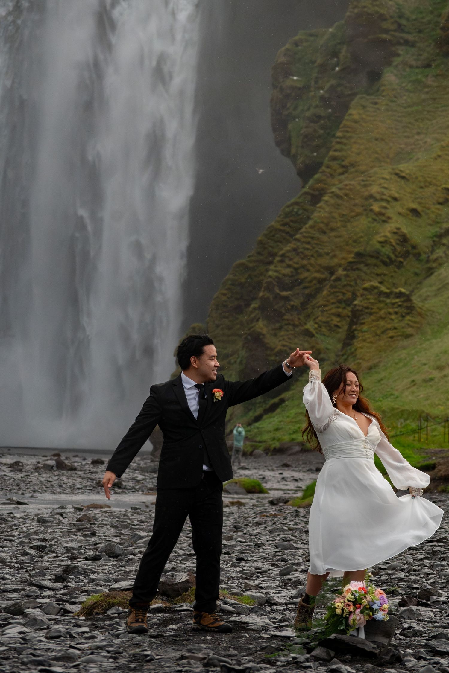 Bride and groom dancing in front of Skogafoss waterfall in Iceland, bride in white dress holding bouquet.