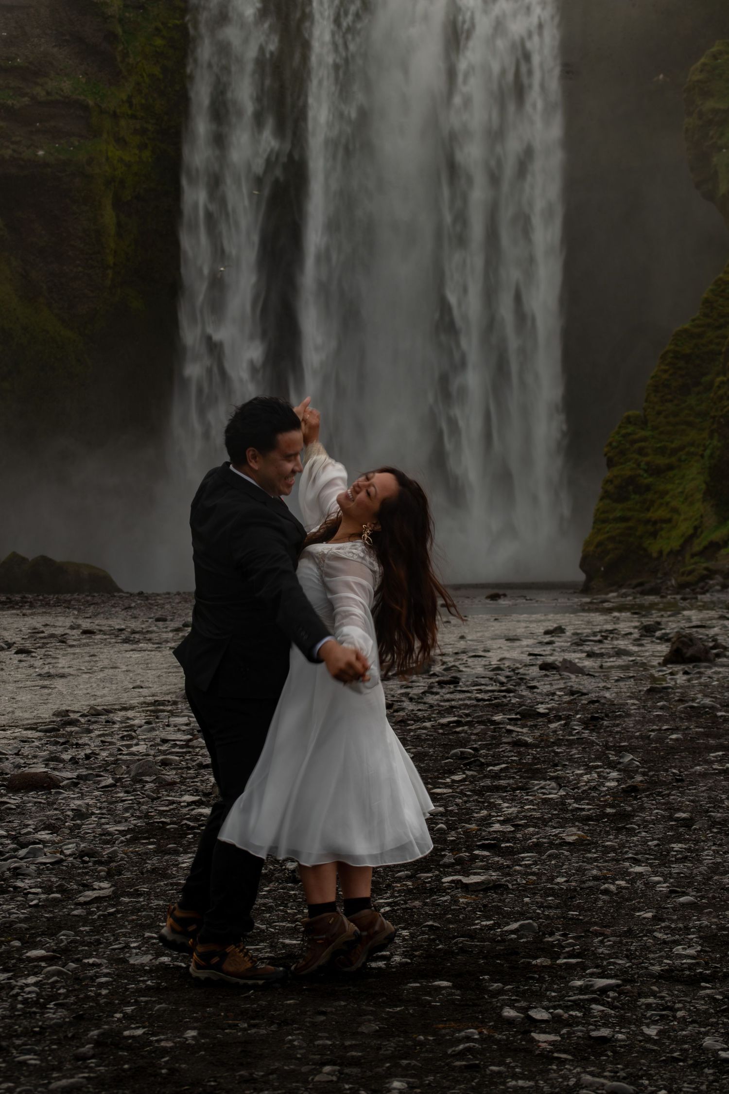 Couple dancing romantically in wedding attire before a majestic waterfall on a rocky landscape.