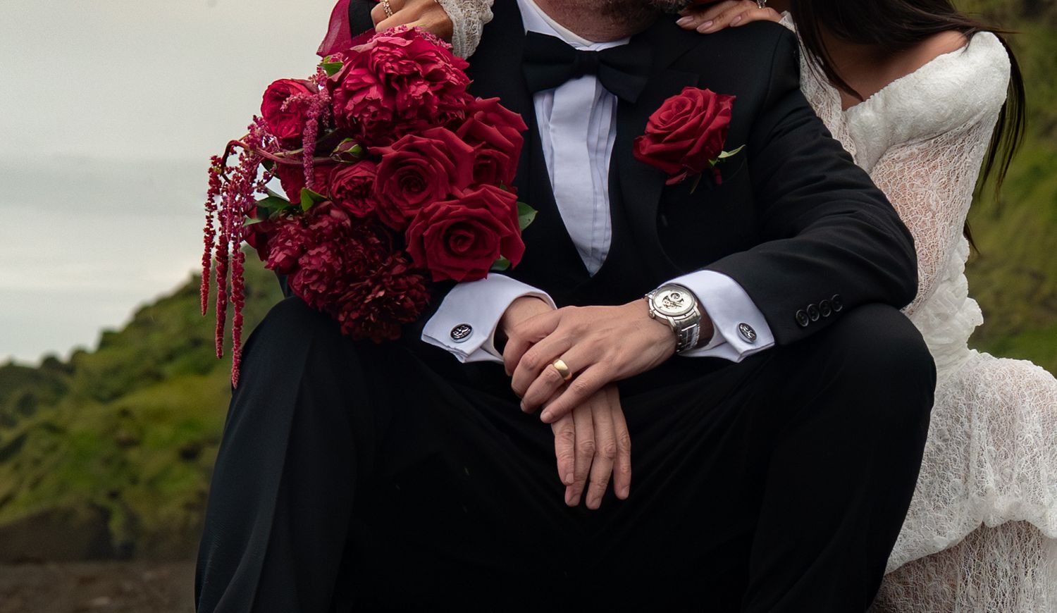 Bride and groom sitting together holding red rose bouquet, groom wearing black tuxedo and silver watch on wedding day.