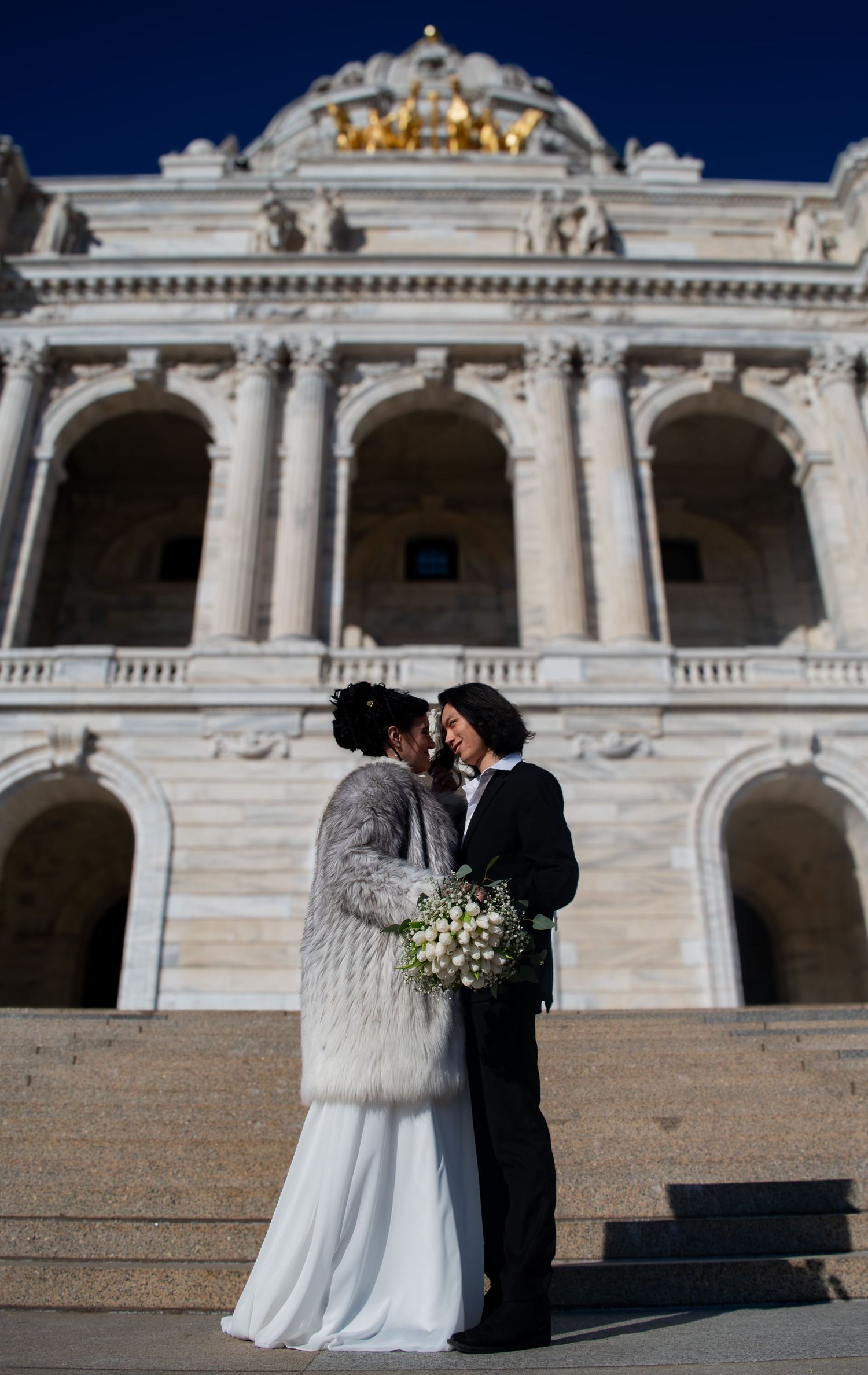Same-sex couple kisses on steps of grand neoclassical capitol building with golden dome backdrop.