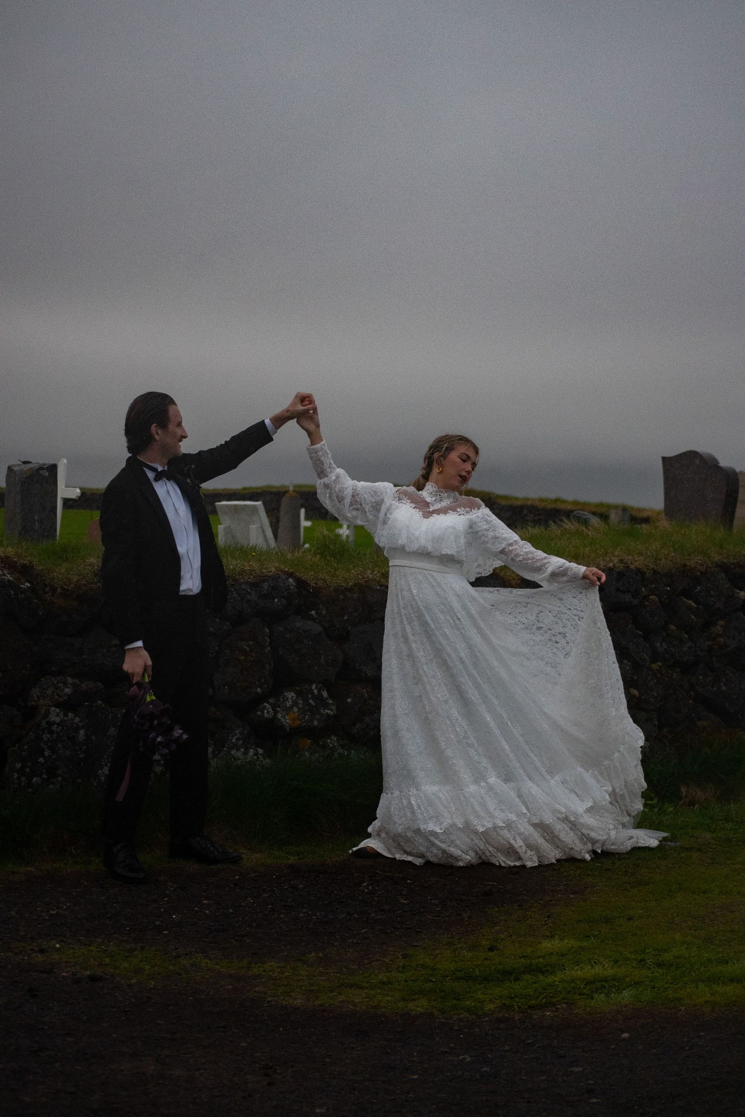 Bride and groom dancing outdoors near stone wall, bride twirling in lace wedding dress under overcast sky.
