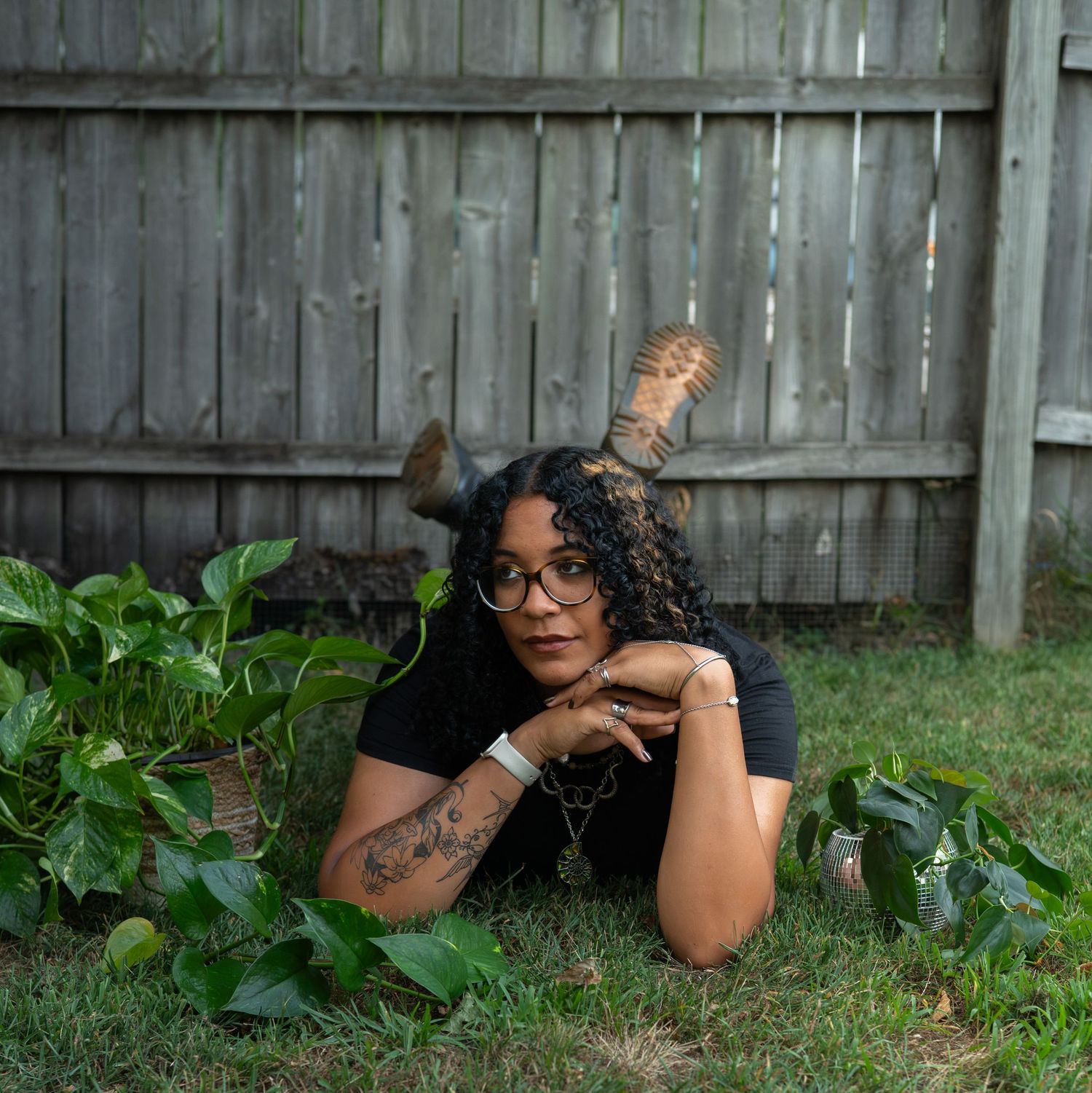 A woman with curly hair and decorative horns lies in green foliage wearing glasses and a black shirt.